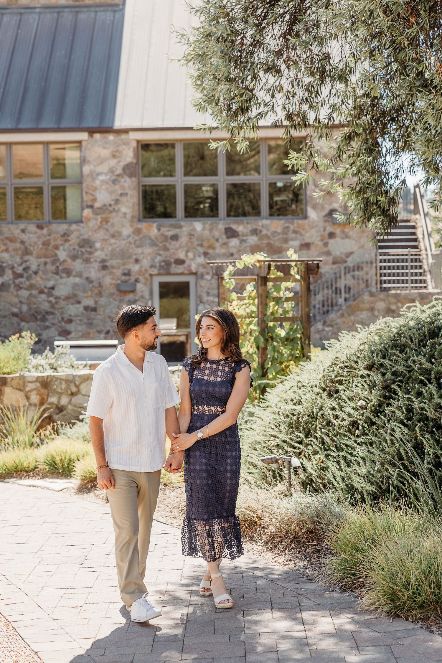 Couple walking along stone pathway beside modern winery building with landscaped gardens.