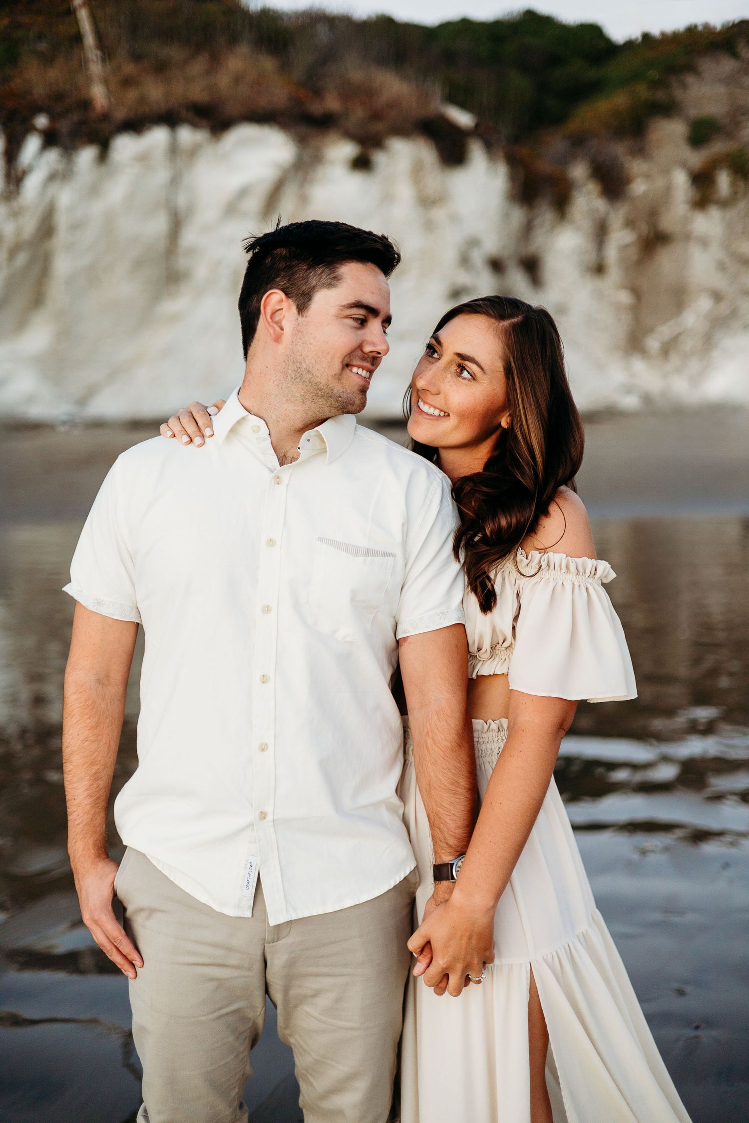 A loving couple shares an intimate moment on the beach with dramatic coastal cliffs in the background.