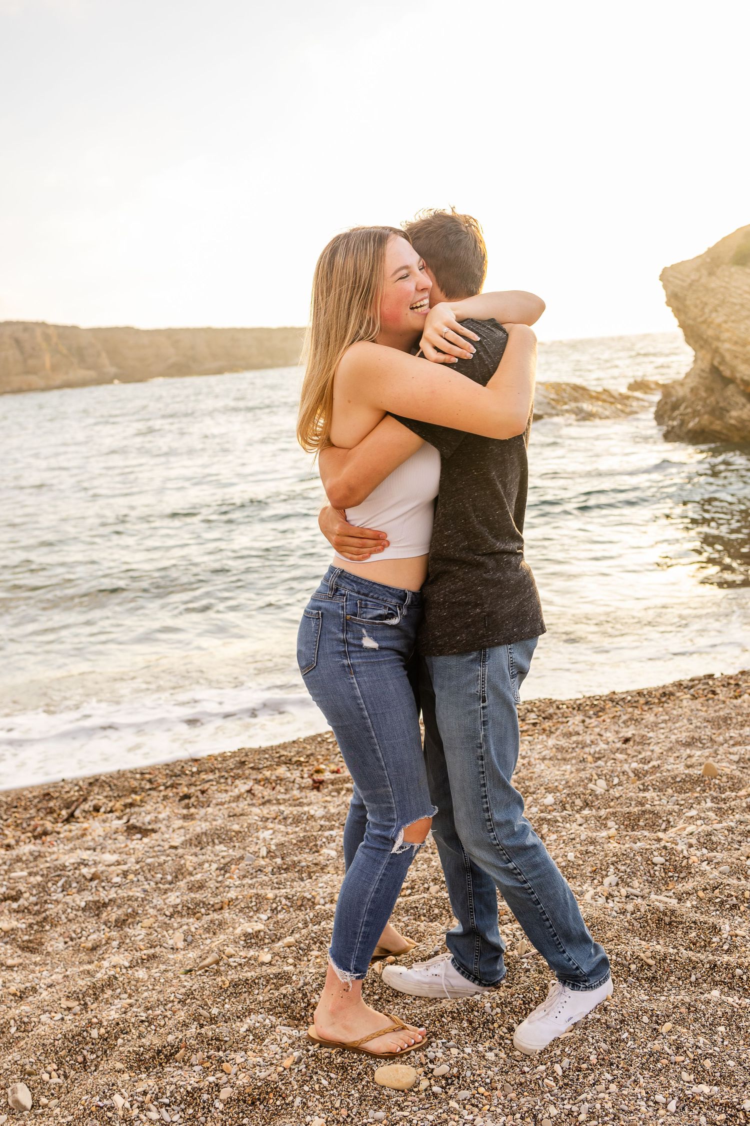 A couple embraces while standing on a rocky beach during sunset.