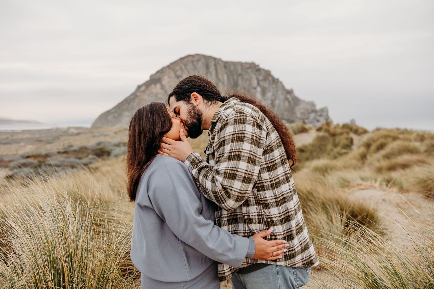 Couple sharing an intimate moment while embracing on a coastal trail with a mountain peak in the background.