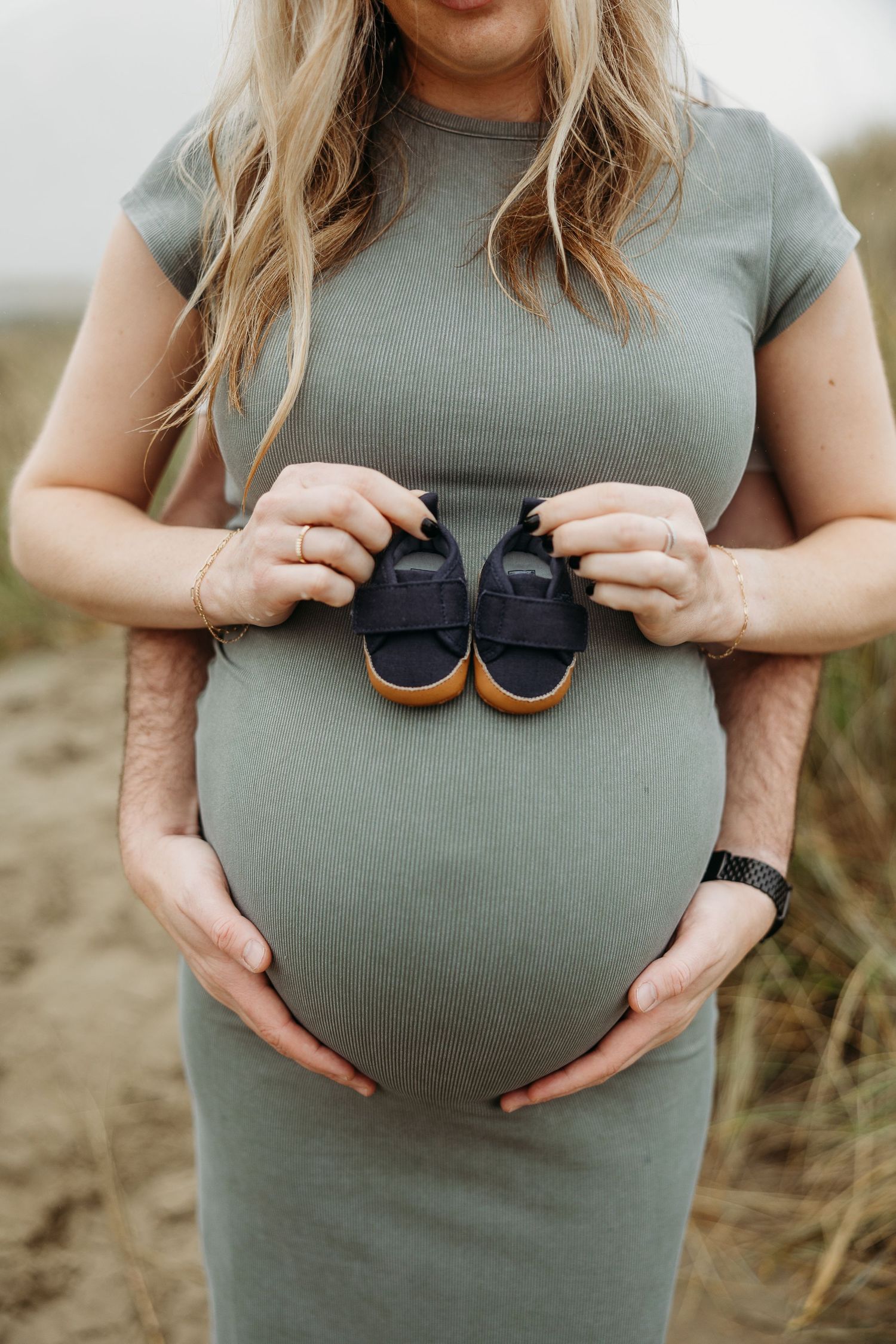 Close-up of pregnant belly with tiny navy baby shoes held against a sage green maternity dress.