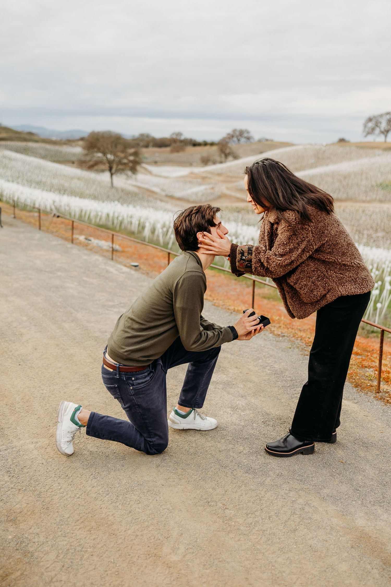 A couple shares a tender moment on a dirt path with rolling hills and vineyards in the background.