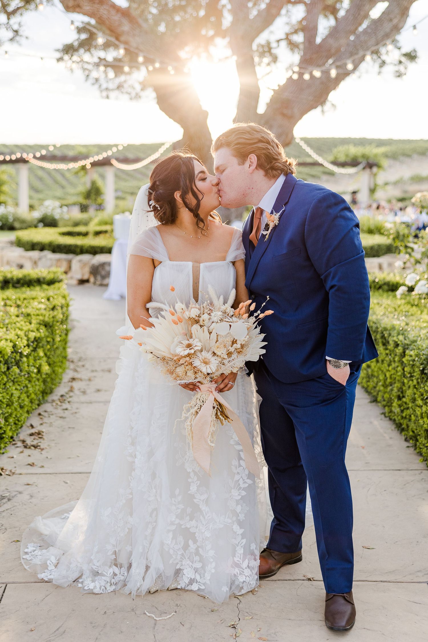 A romantic wedding couple share a kiss in a garden setting with beautiful sunlight streaming through trees.