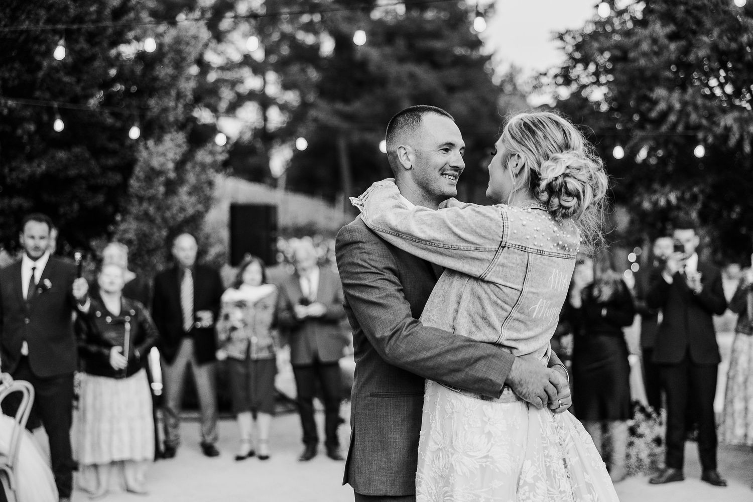 Black and white photo of couple dancing and laughing at outdoor reception under string lights.
