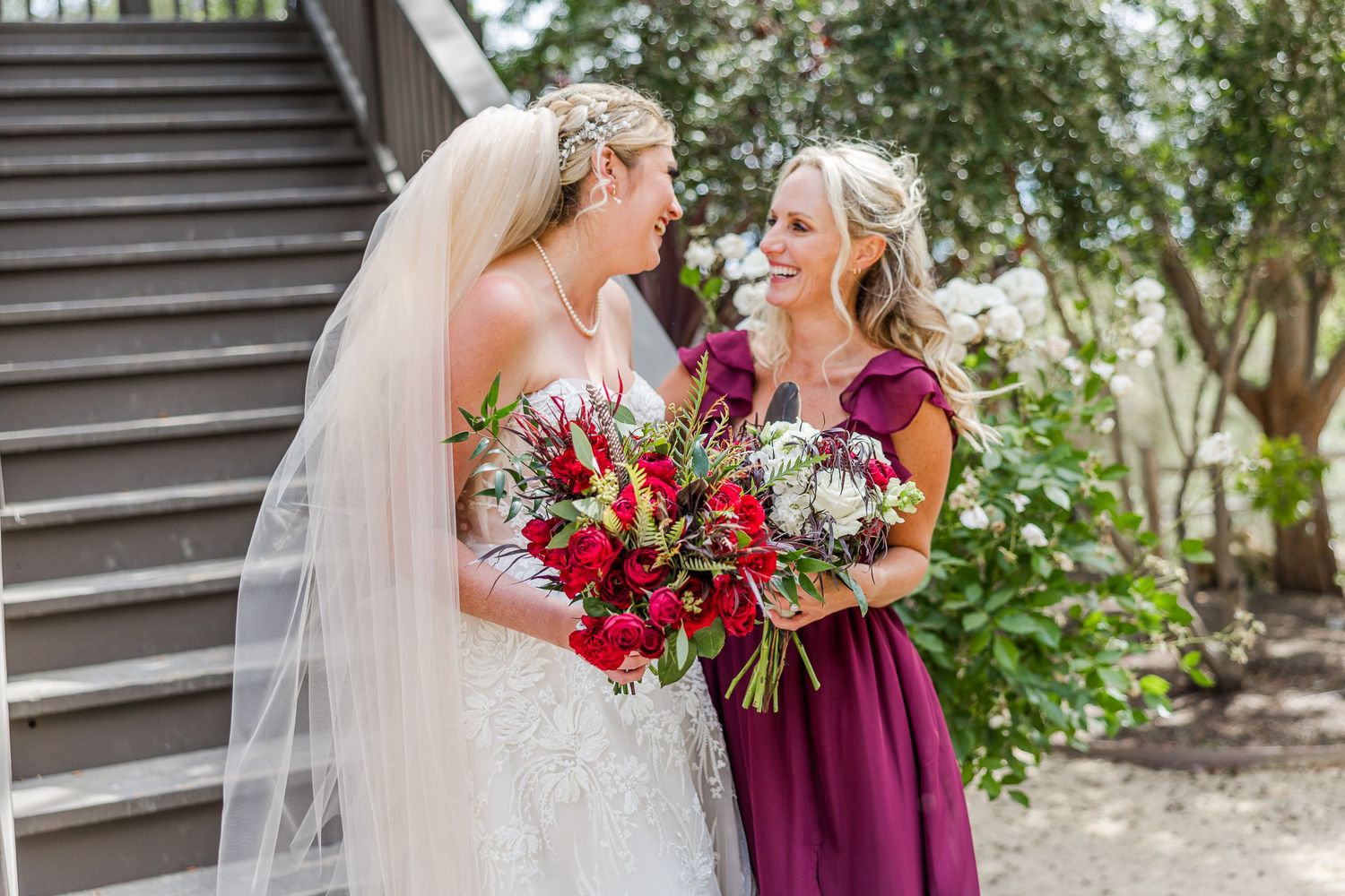A bride holds a stunning red bouquet while sharing a joyful moment outdoors.
