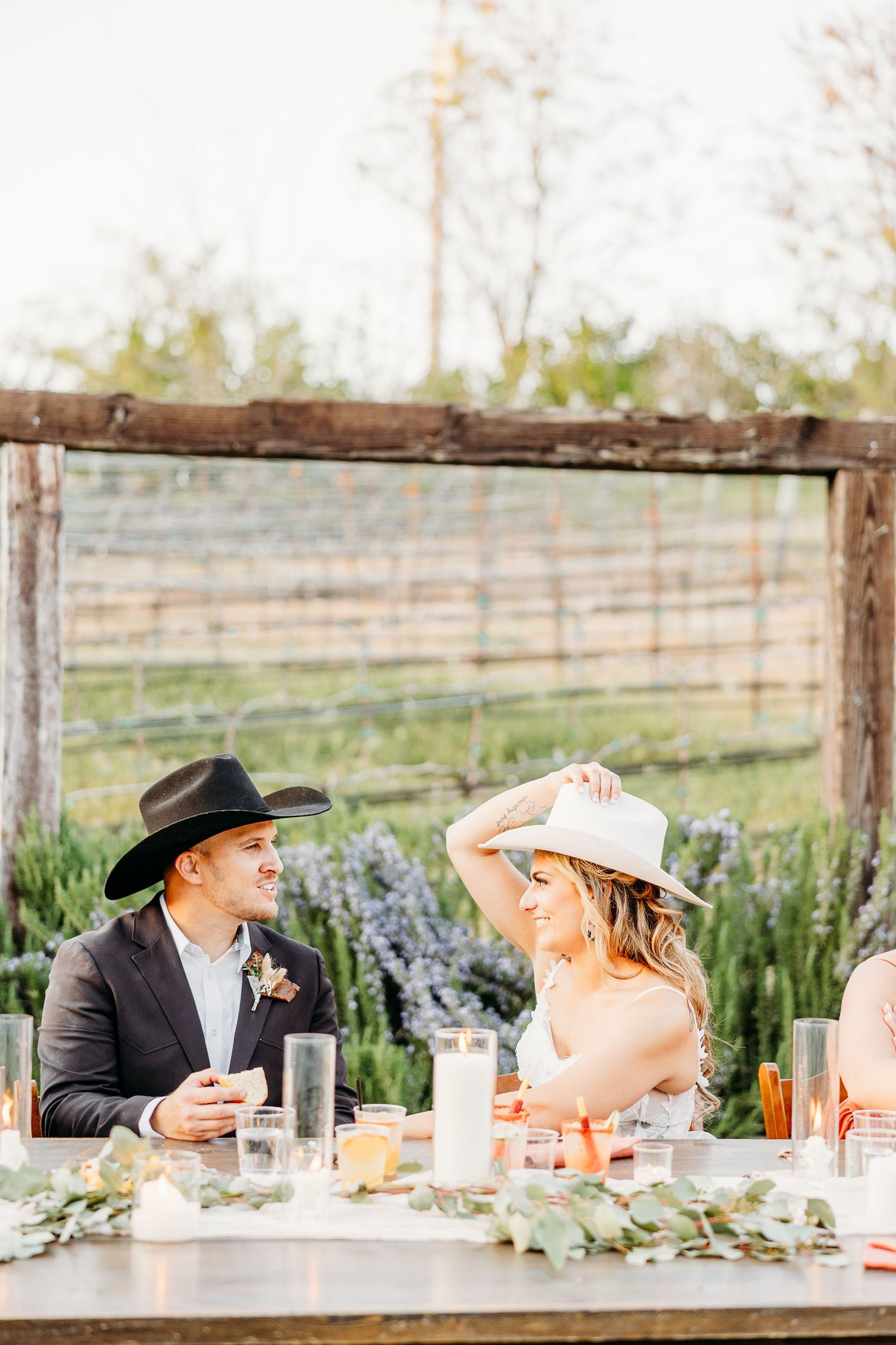 A couple in western wedding attire sharing a candid moment at an outdoor table with rustic decor.