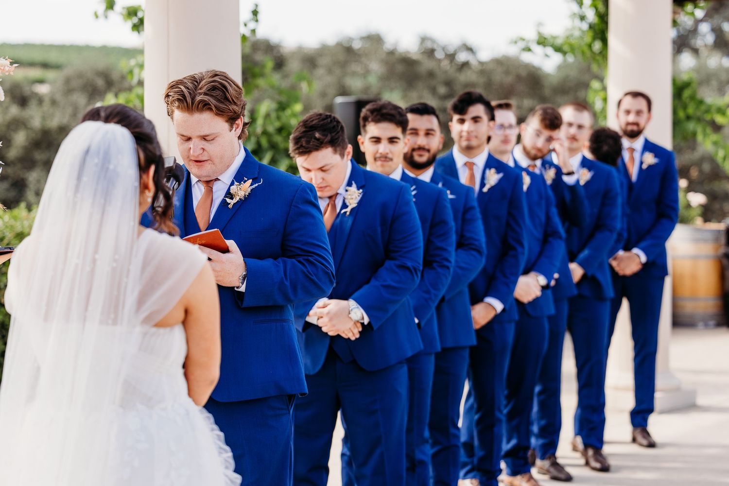 Wedding party members in vibrant blue suits line up during an outdoor ceremony.