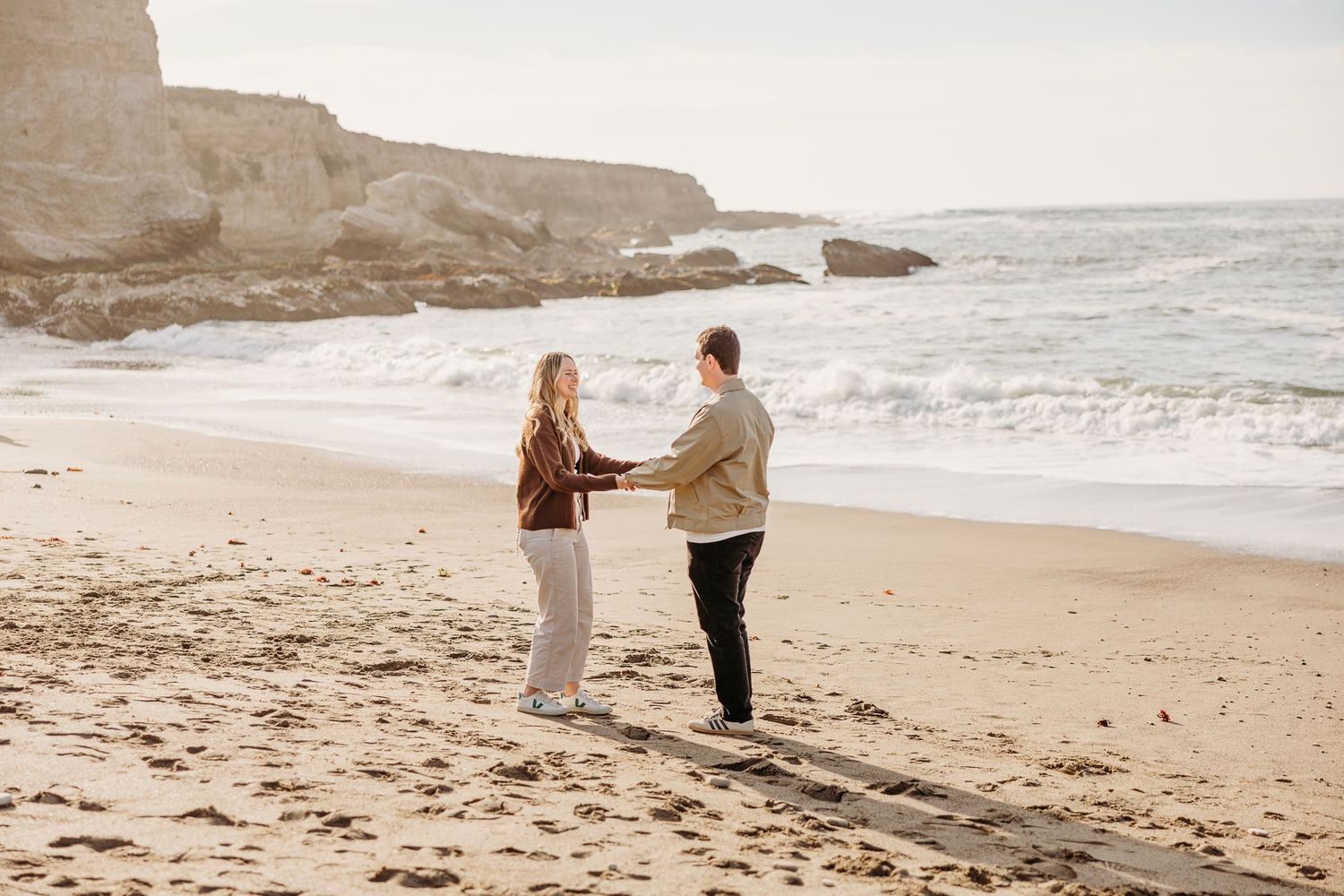 A couple stands together holding hands on a scenic beach with dramatic cliffs in the background.