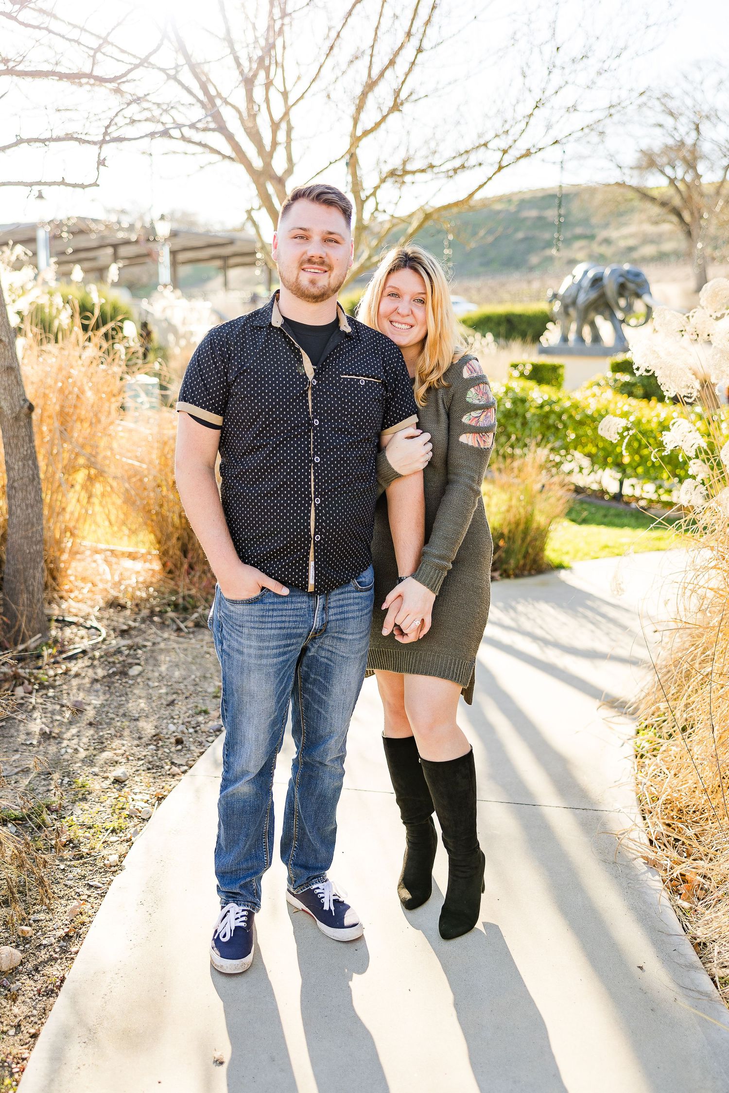 A couple stands together on a concrete path with dried grasses and hills in the background.