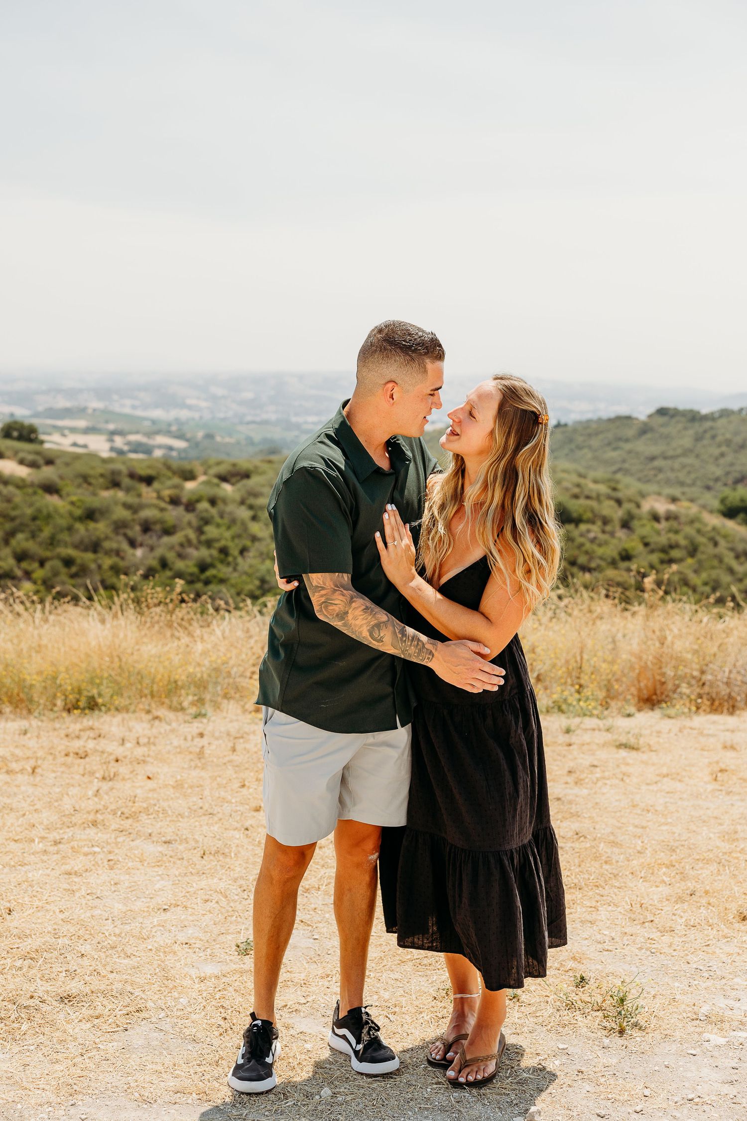 A couple celebrates their engagement on a hilltop overlooking a scenic valley landscape.