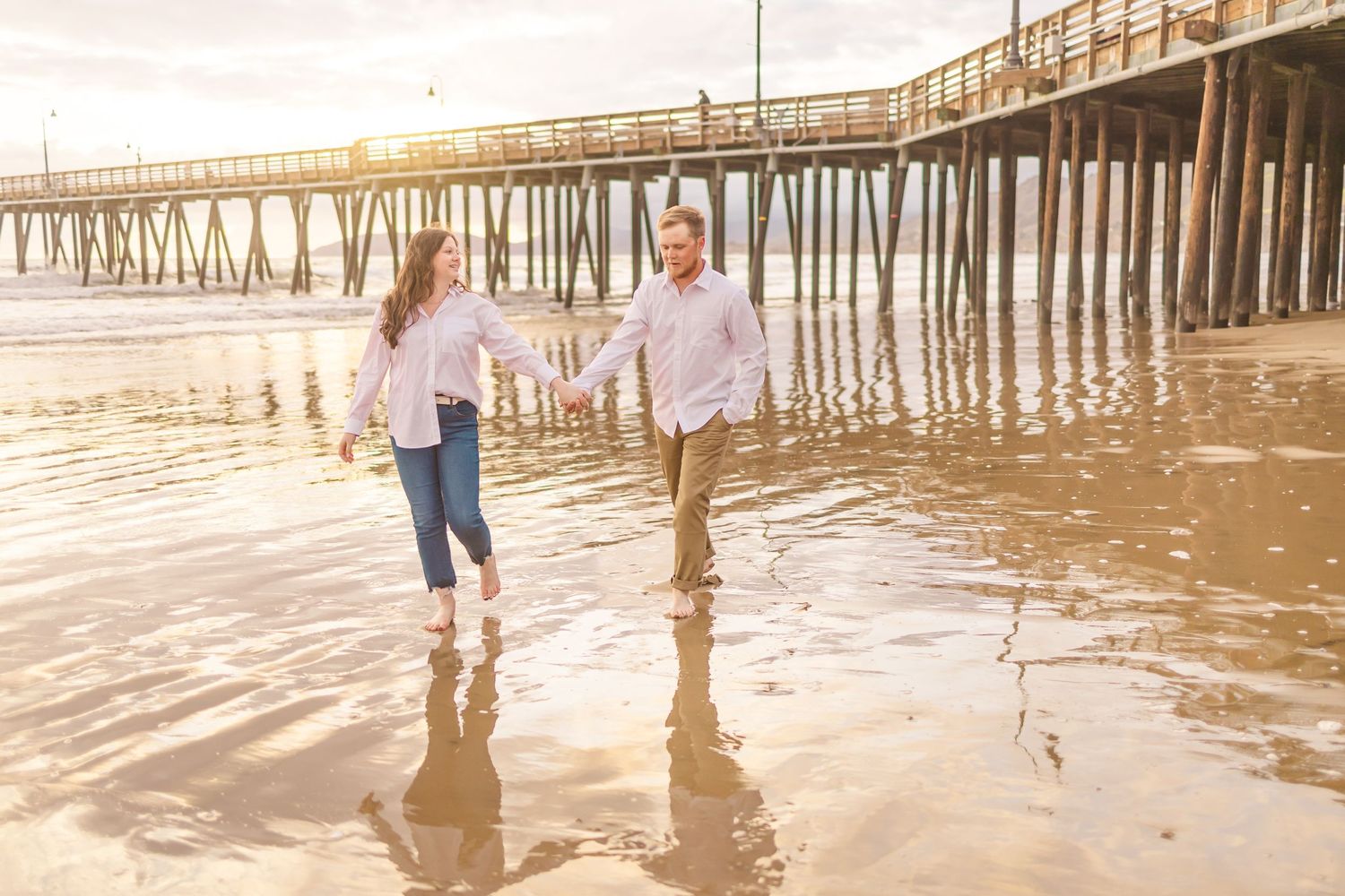 Couple splashes in shallow water near pier during sunset beach photoshoot.