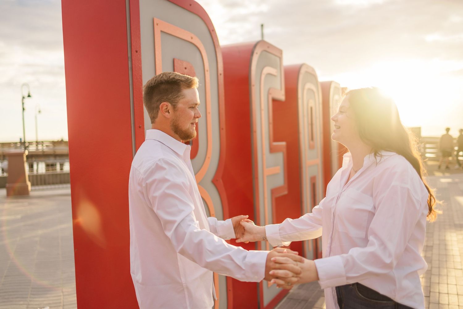 Couple holds hands and shares a romantic moment in front of large red letters during golden hour.