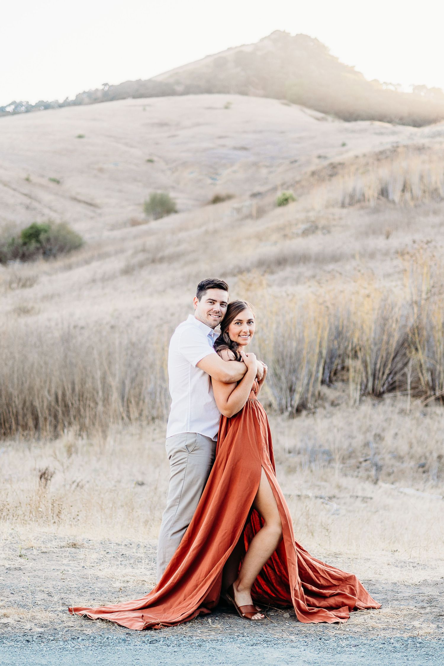 A couple poses dramatically against rolling hills, with the person in a flowing rust-colored dress.