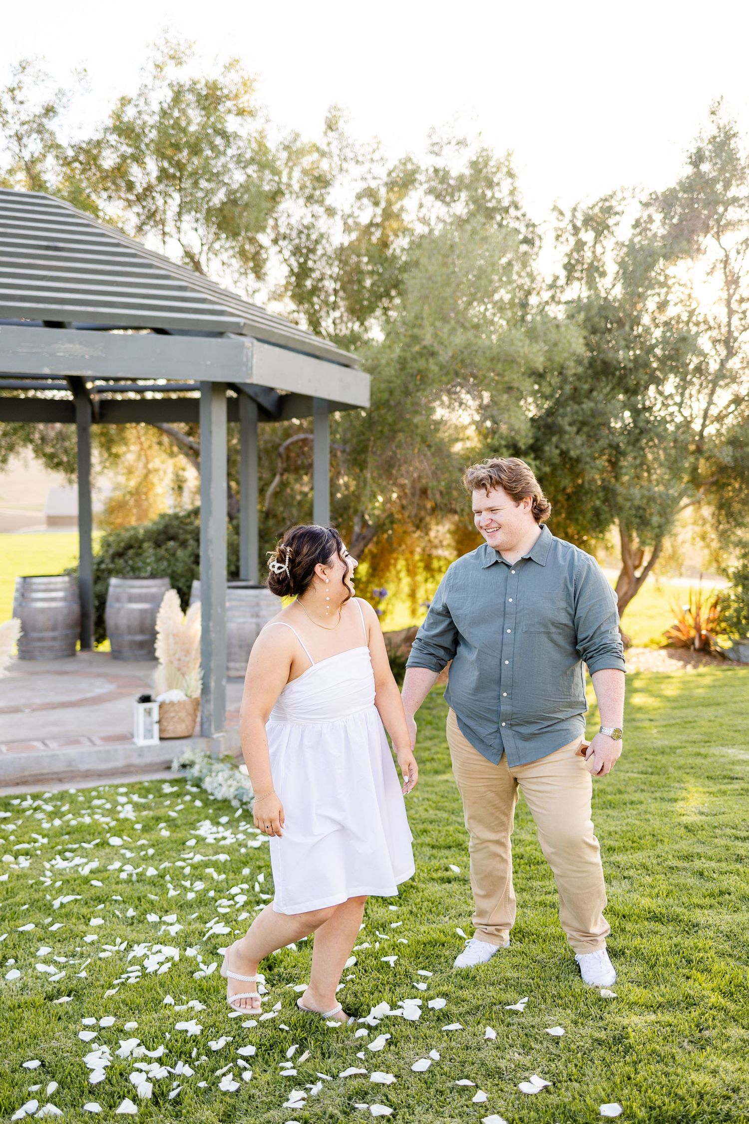 A couple walks together on grass strewn with white flower petals near a rustic gazebo.