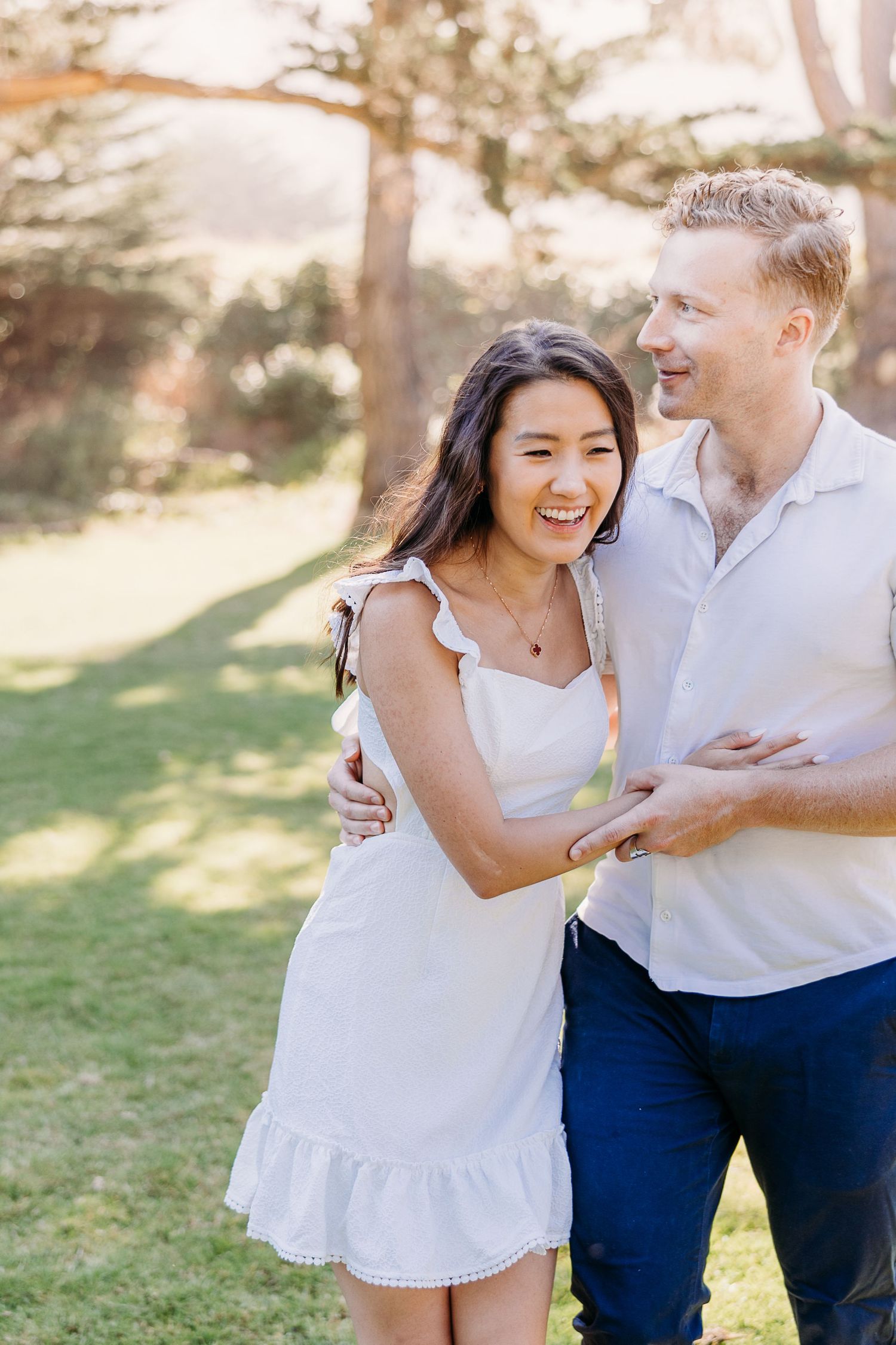 A romantic couple laughs together in a sunlit garden setting wearing coordinated white and blue outfits.
