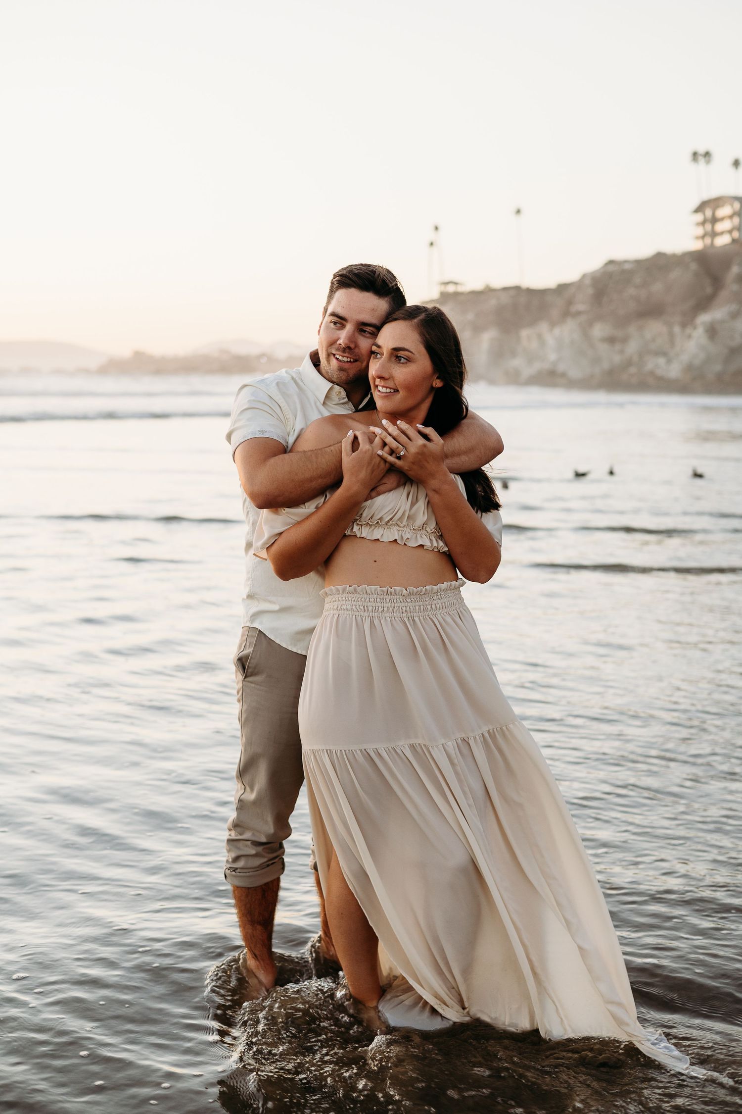 Happy couple embracing on rocky beach with cliffs and ocean waves at sunset.
