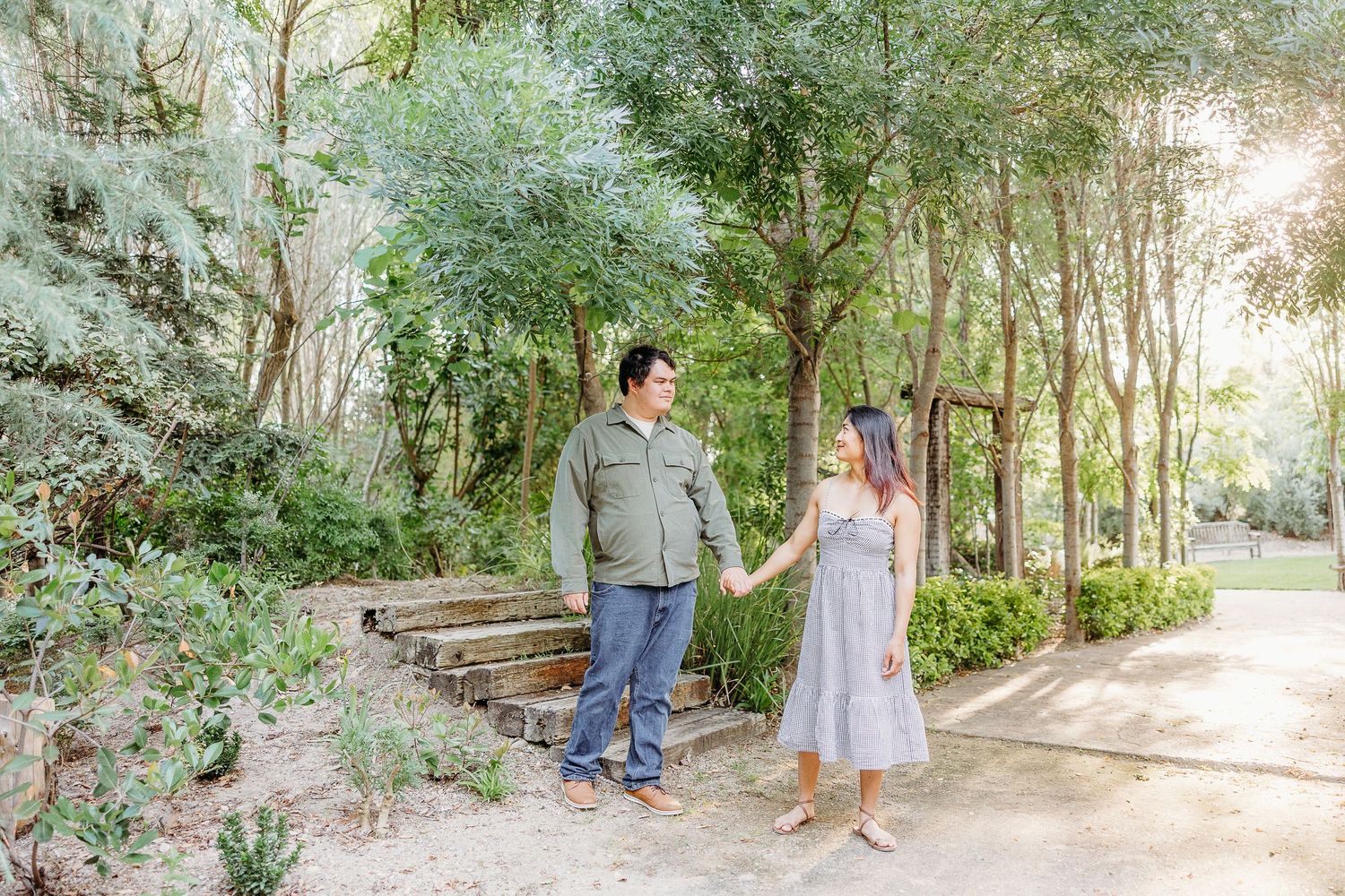 A couple holds hands while standing on a garden path surrounded by lush greenery and trees in soft sunlight.
