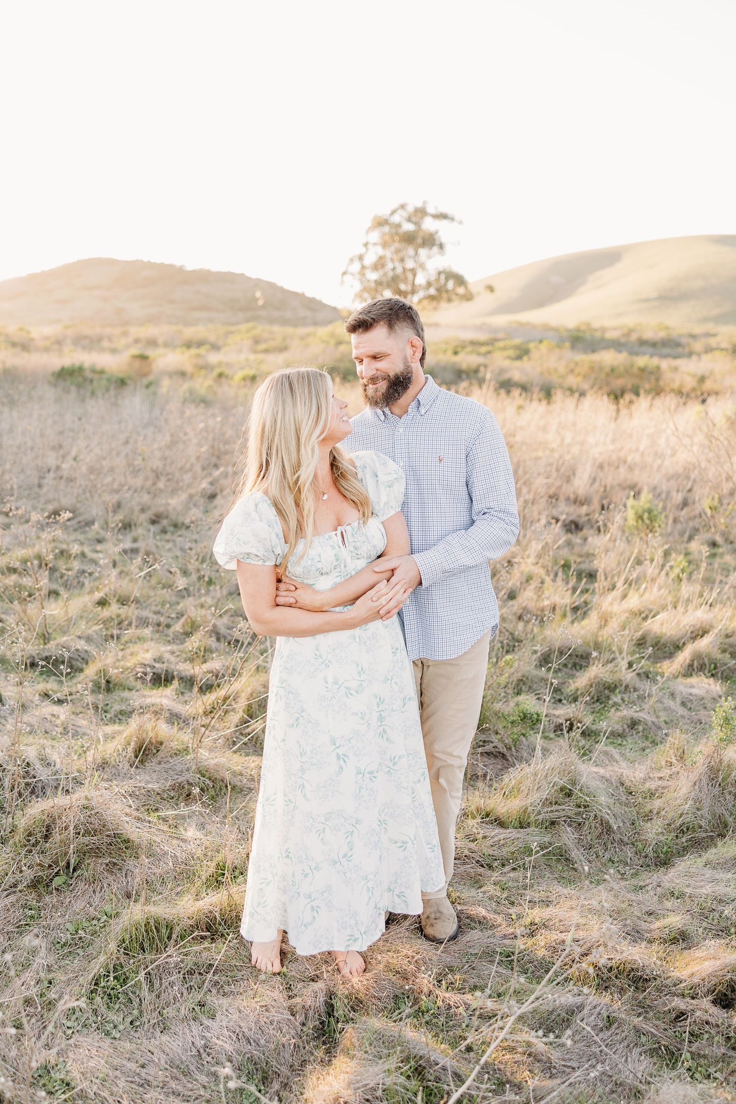 A loving couple embraces on a rolling hillside at sunset in soft earthy tones.