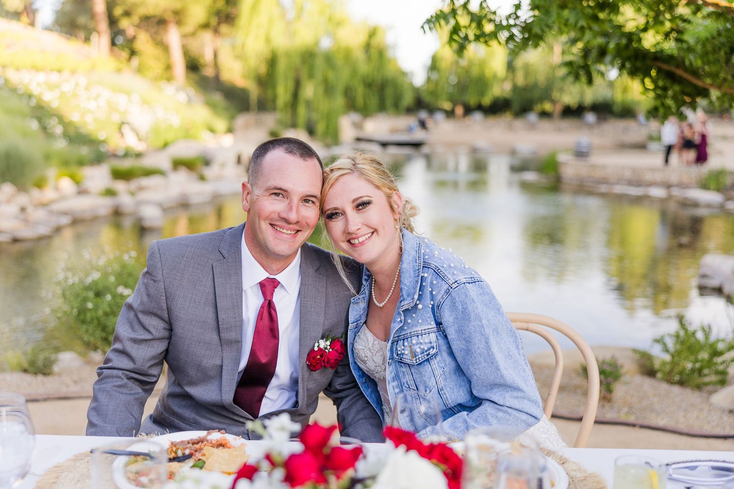 A couple sitting at a table by a scenic pond with red floral centerpieces.