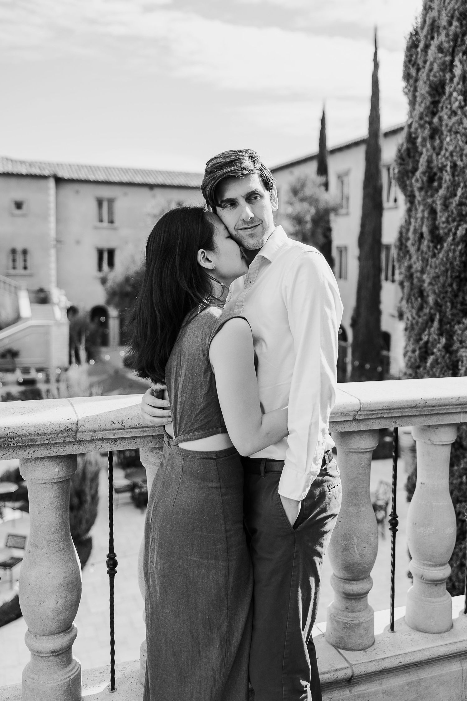A romantic black and white couple portrait on a balcony with stone balusters and cypress trees in the background.