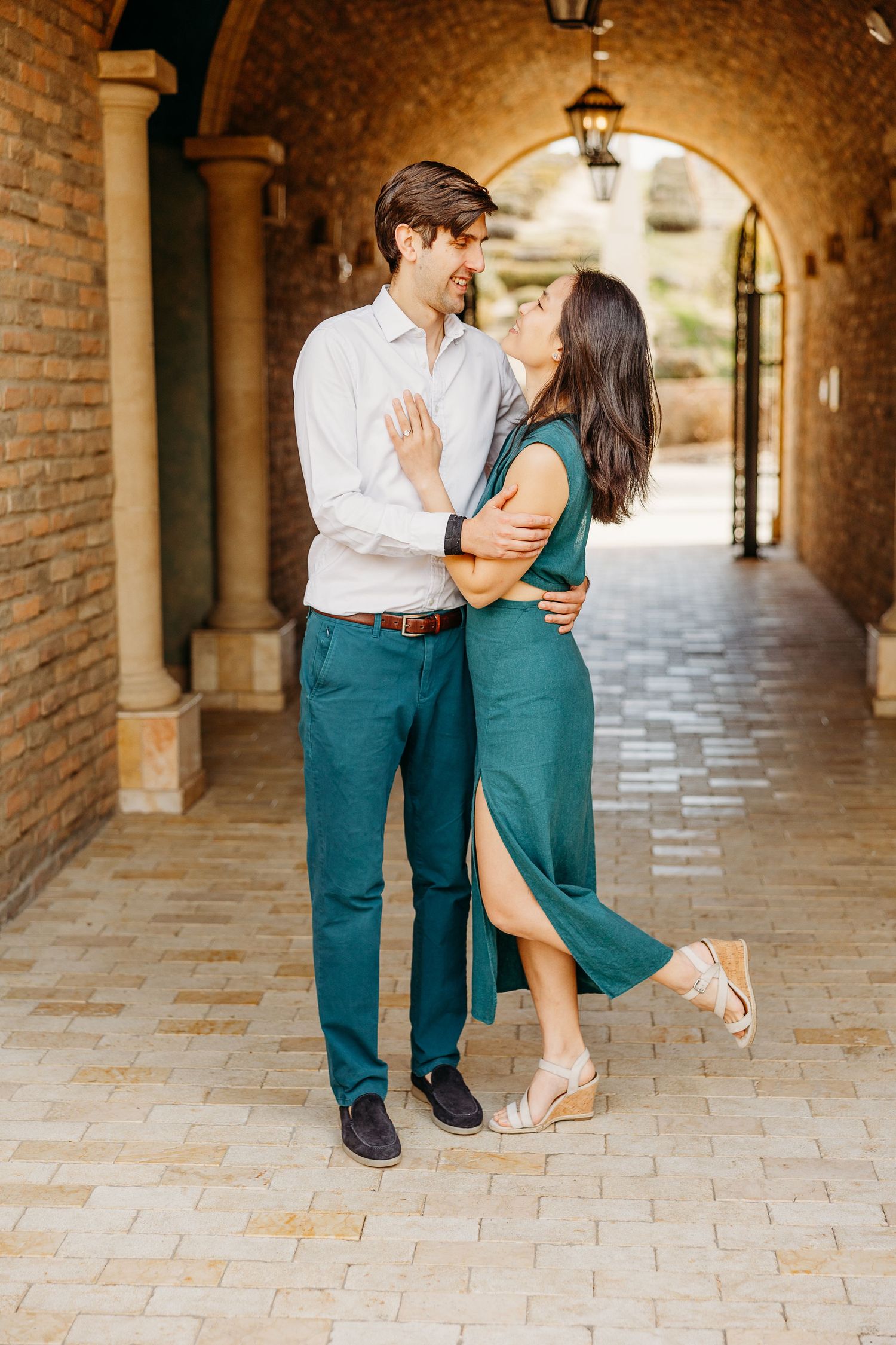Romantic couple in coordinating green outfits embrace in arched brick hallway with vintage lantern lighting.