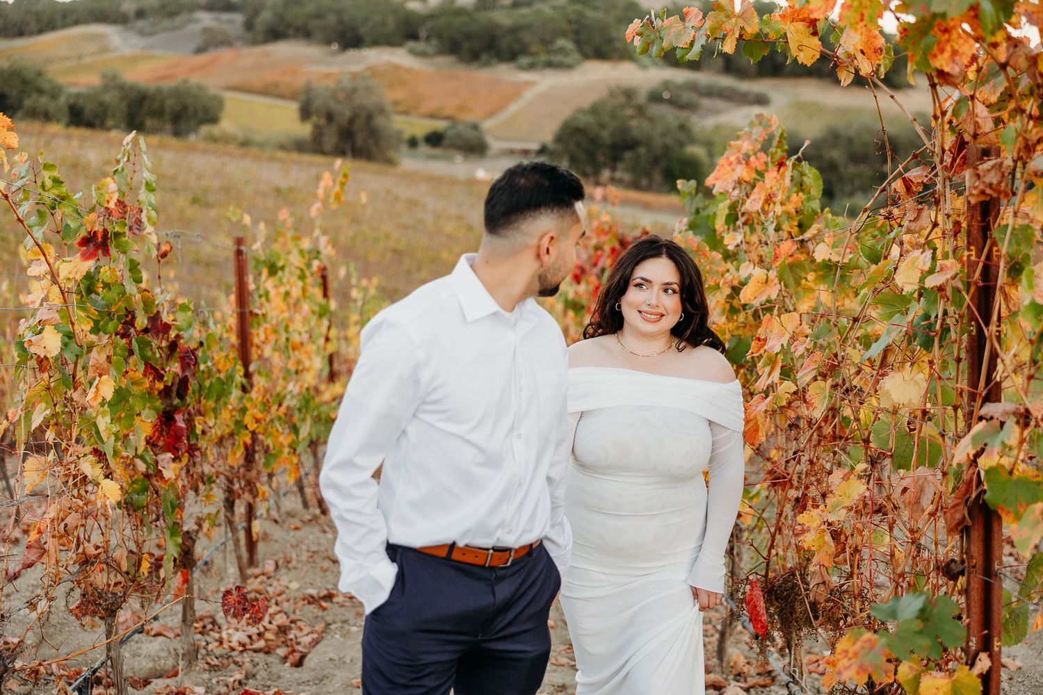 Couple poses together in vineyard with autumn colored leaves and rolling hills in background.