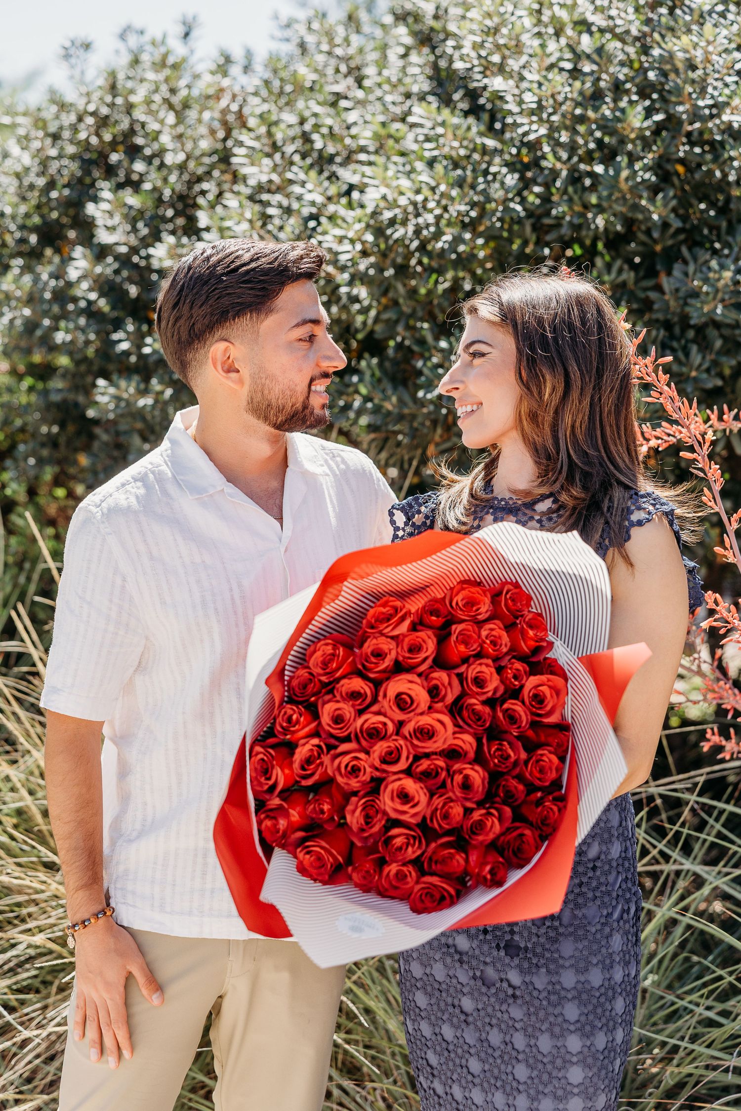 A couple shares a romantic moment in a vineyard while holding a large bouquet of red roses.