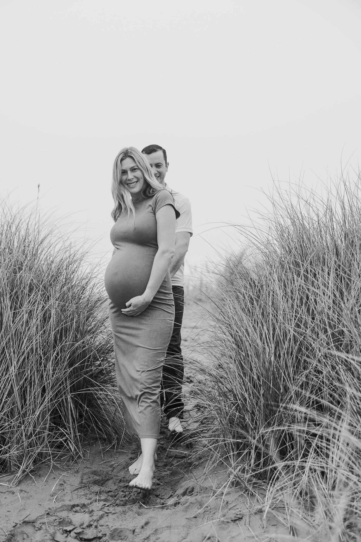 Black and white maternity photo of a couple standing among beach grass on sandy dunes.