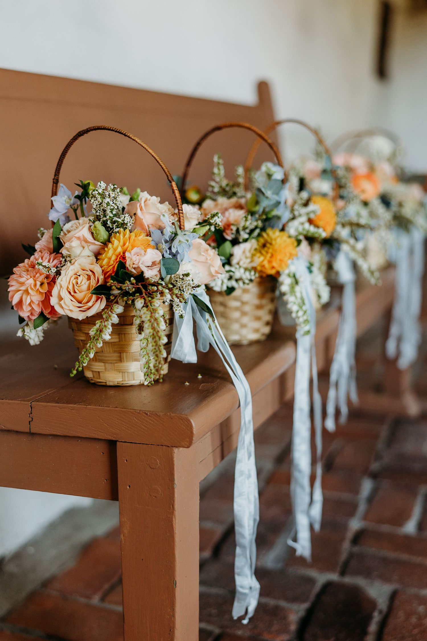 Floral centerpieces with roses and dahlias in woven baskets displayed on brown wooden benches with trailing ribbons.