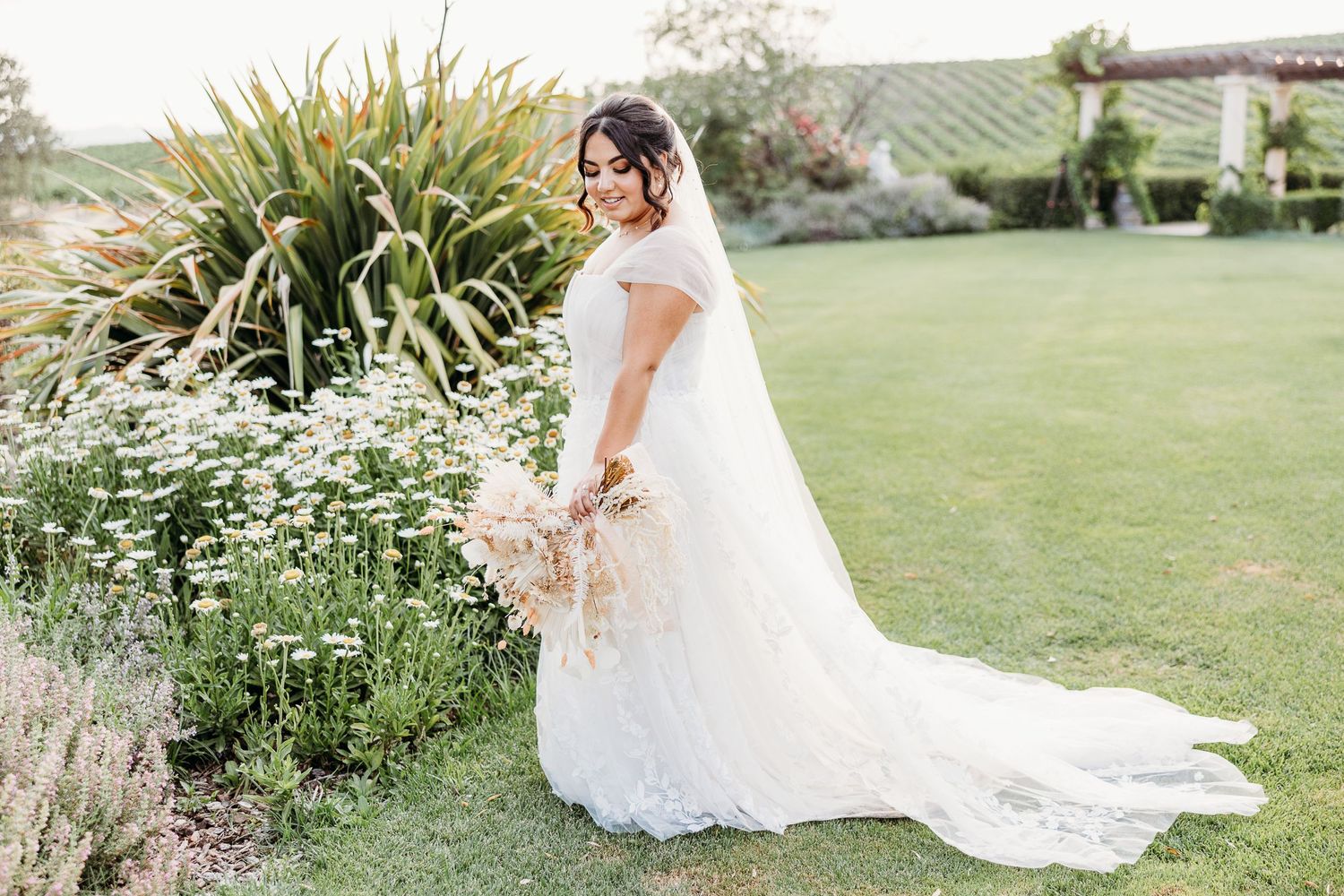 Full length portrait of bride in flowing wedding gown with dried floral bouquet in garden.