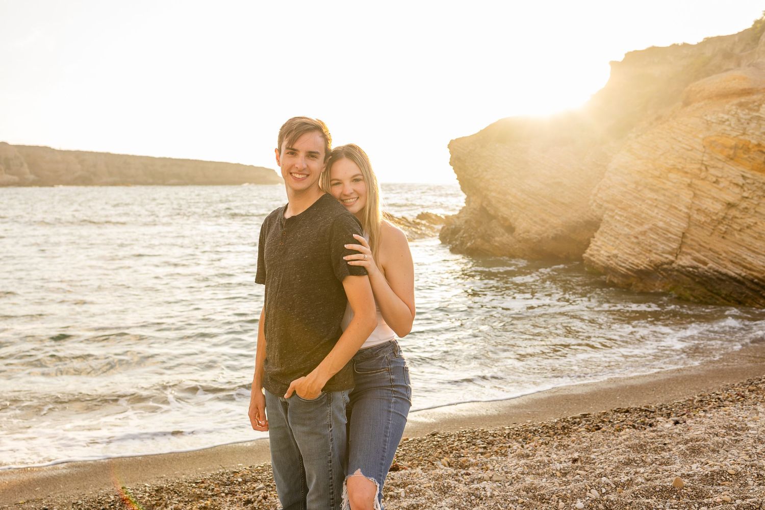 A couple shares a joyful moment together on a rocky beach during golden hour.
