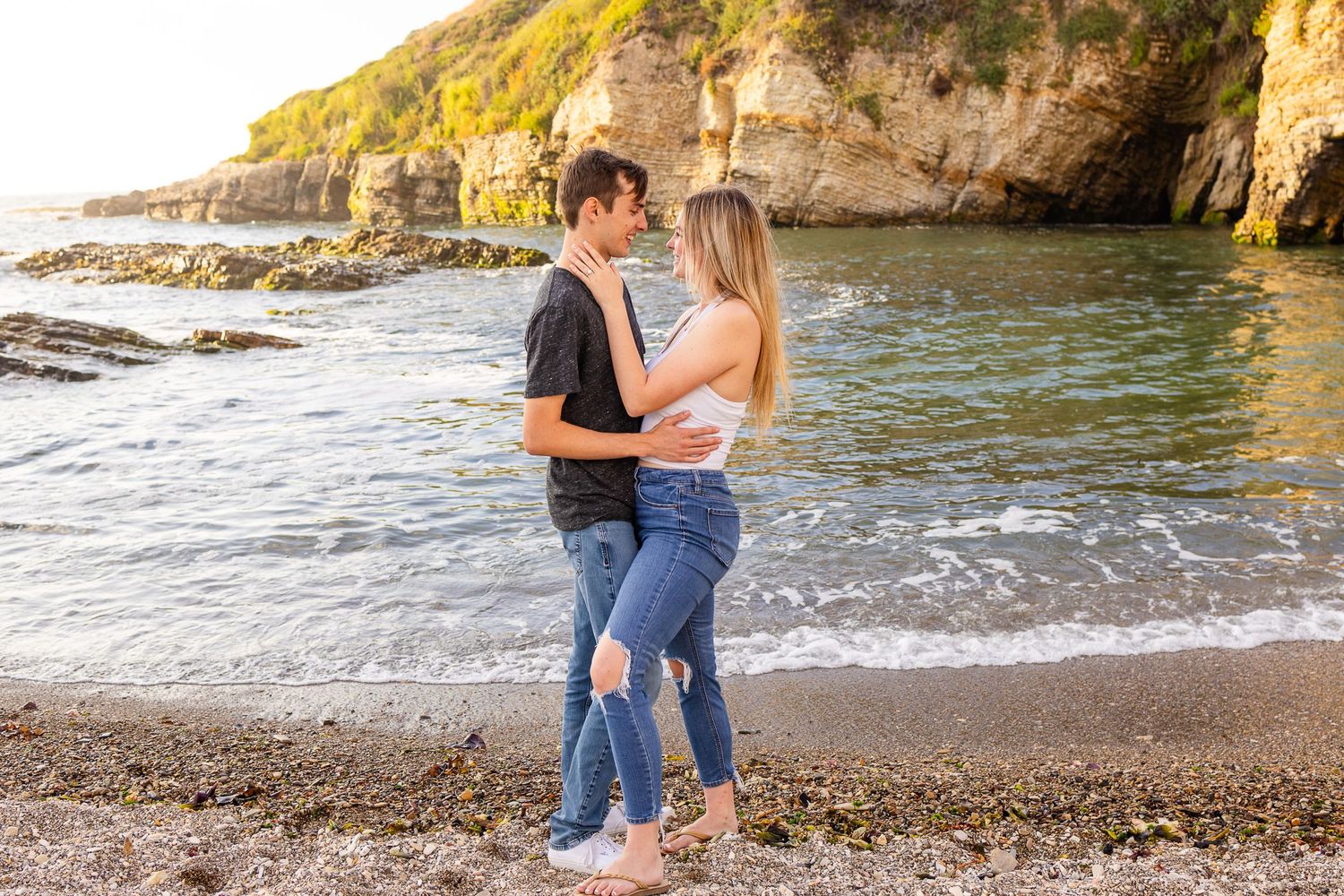 A couple embraces on a scenic beach with rocky cliffs in the background during golden hour.