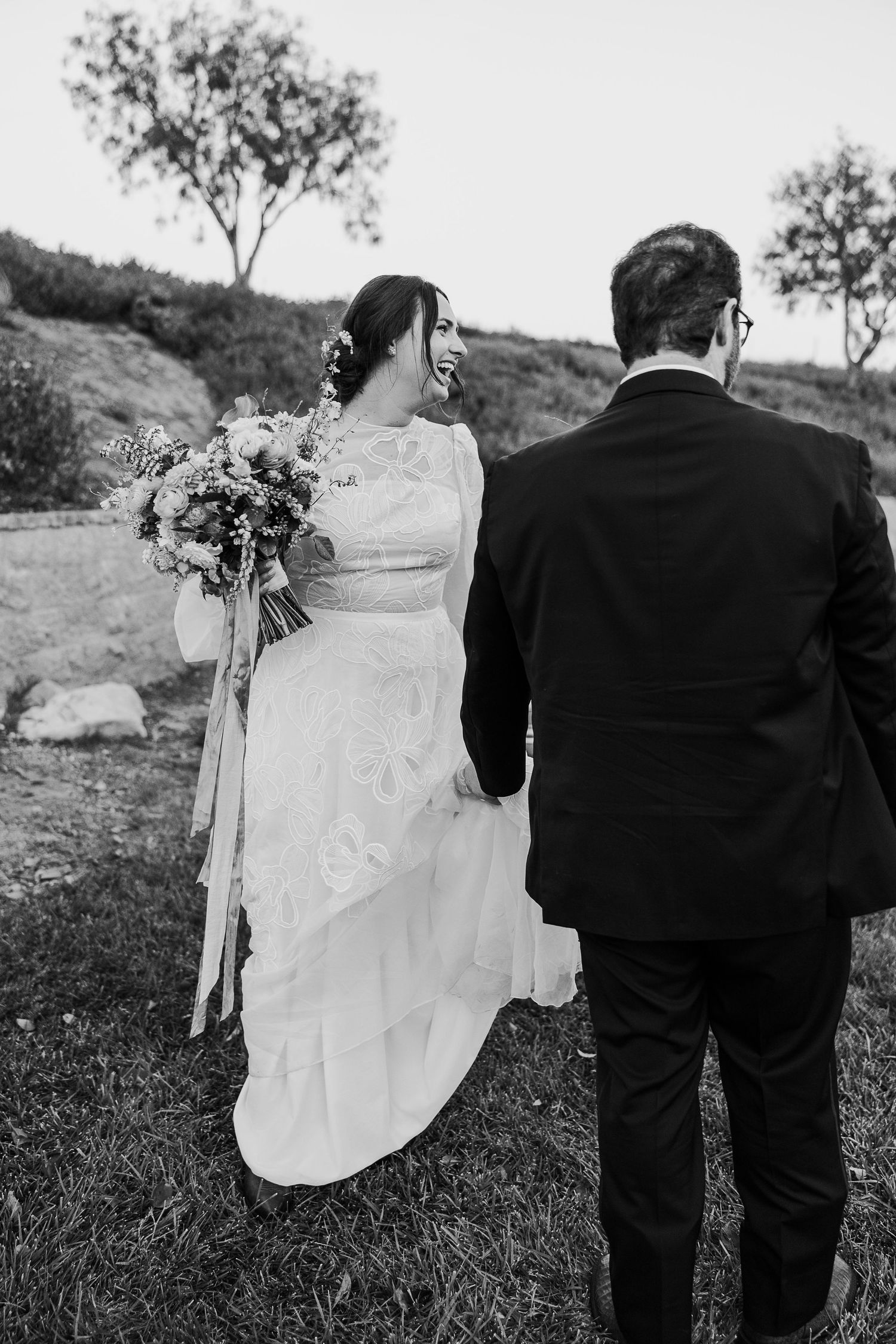 A black and white portrait of newlyweds from behind, holding bouquet against hillside scenery.