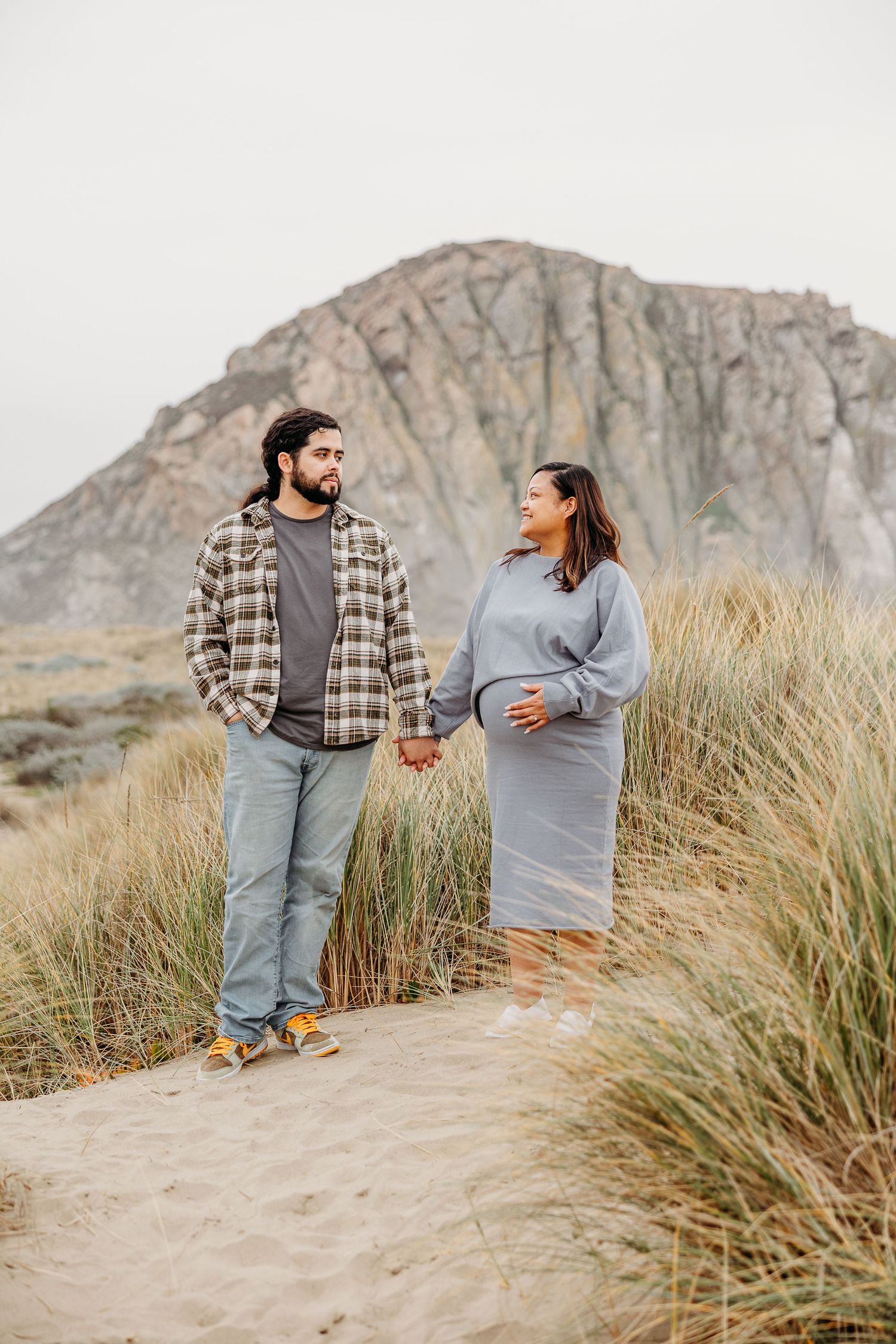 A couple holding hands while standing in coastal dune grass with a rocky cliff behind them.
