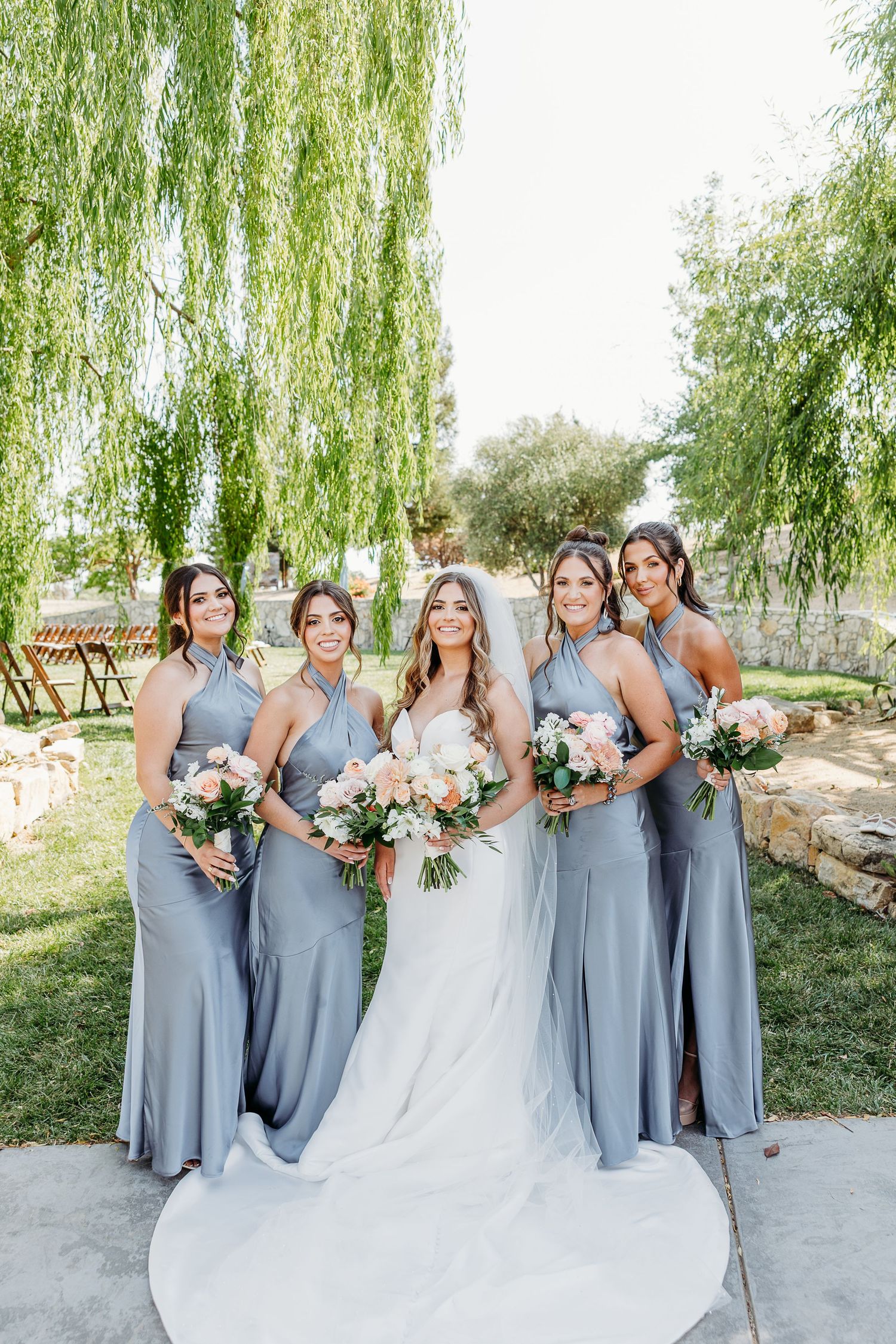 Bridal party in grey dresses poses elegantly under a willow tree at an outdoor wedding.