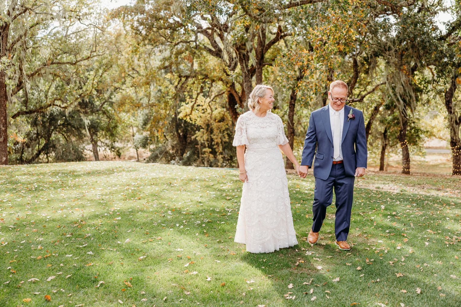 Couple walks hand in hand through a scenic garden setting with moss-draped oak trees.