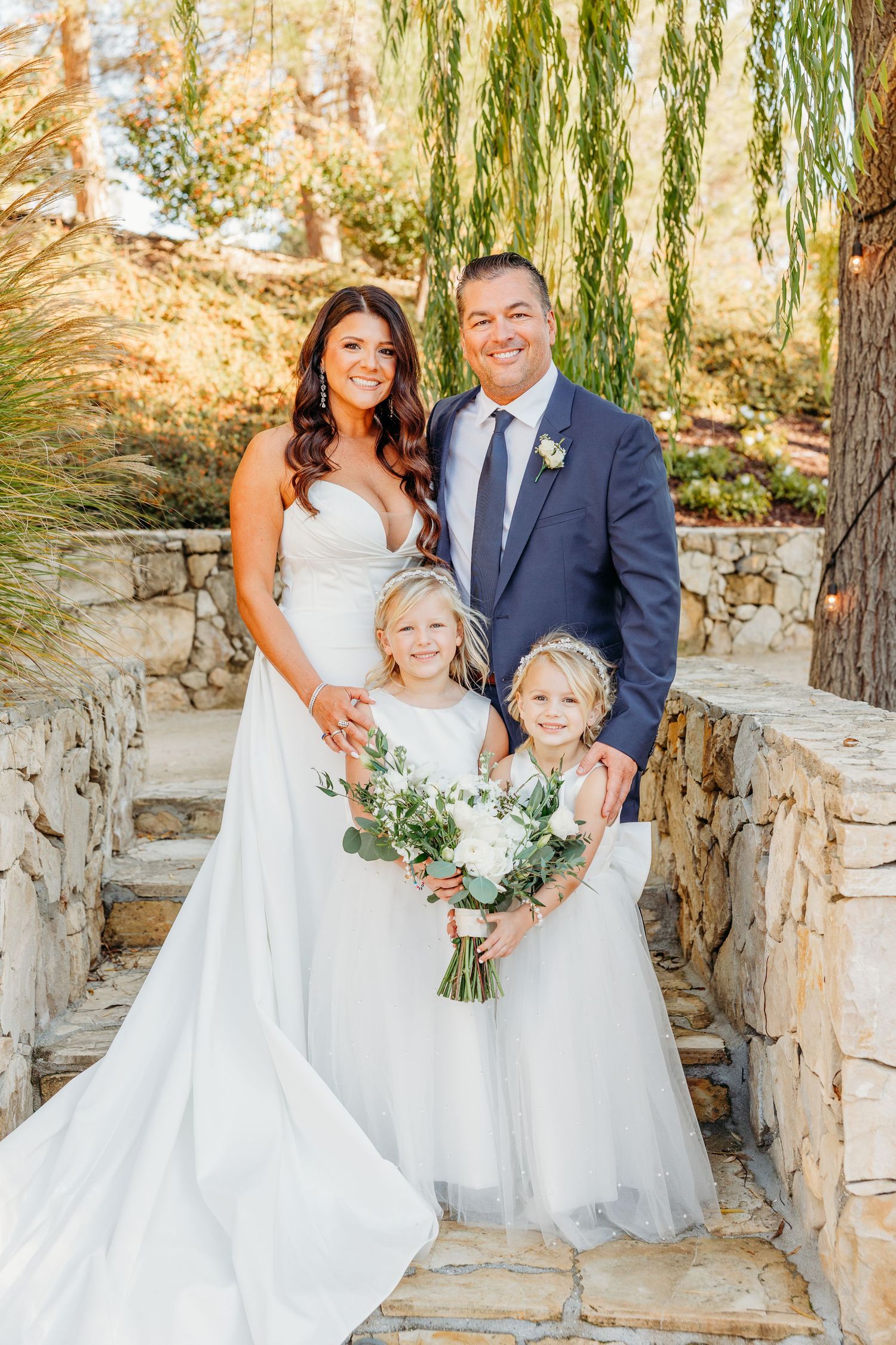 A wedding couple poses with two young flower girls in white dresses on stone steps under willow trees.