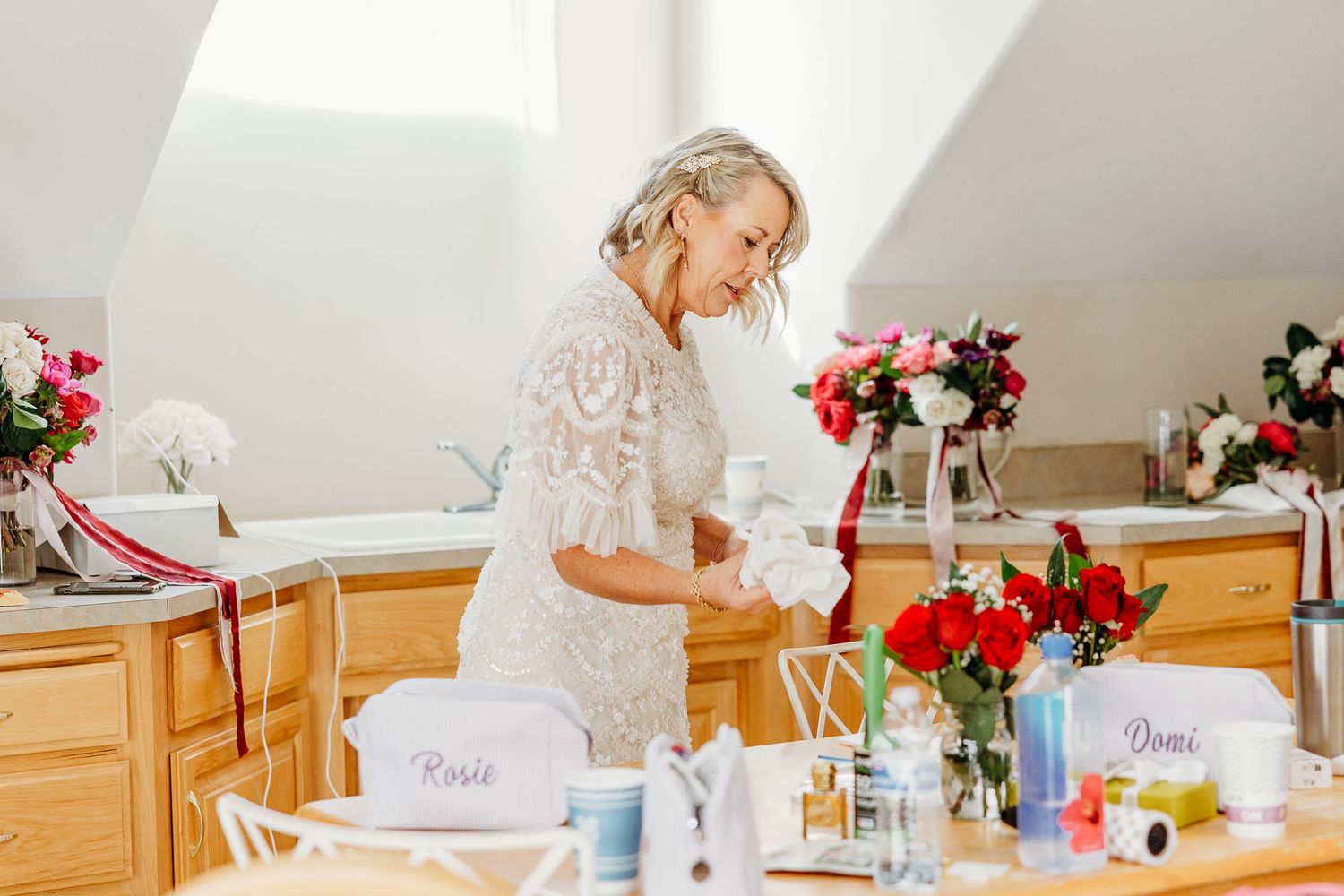 Preparation scene with white lace dress and red floral arrangements in bright room.