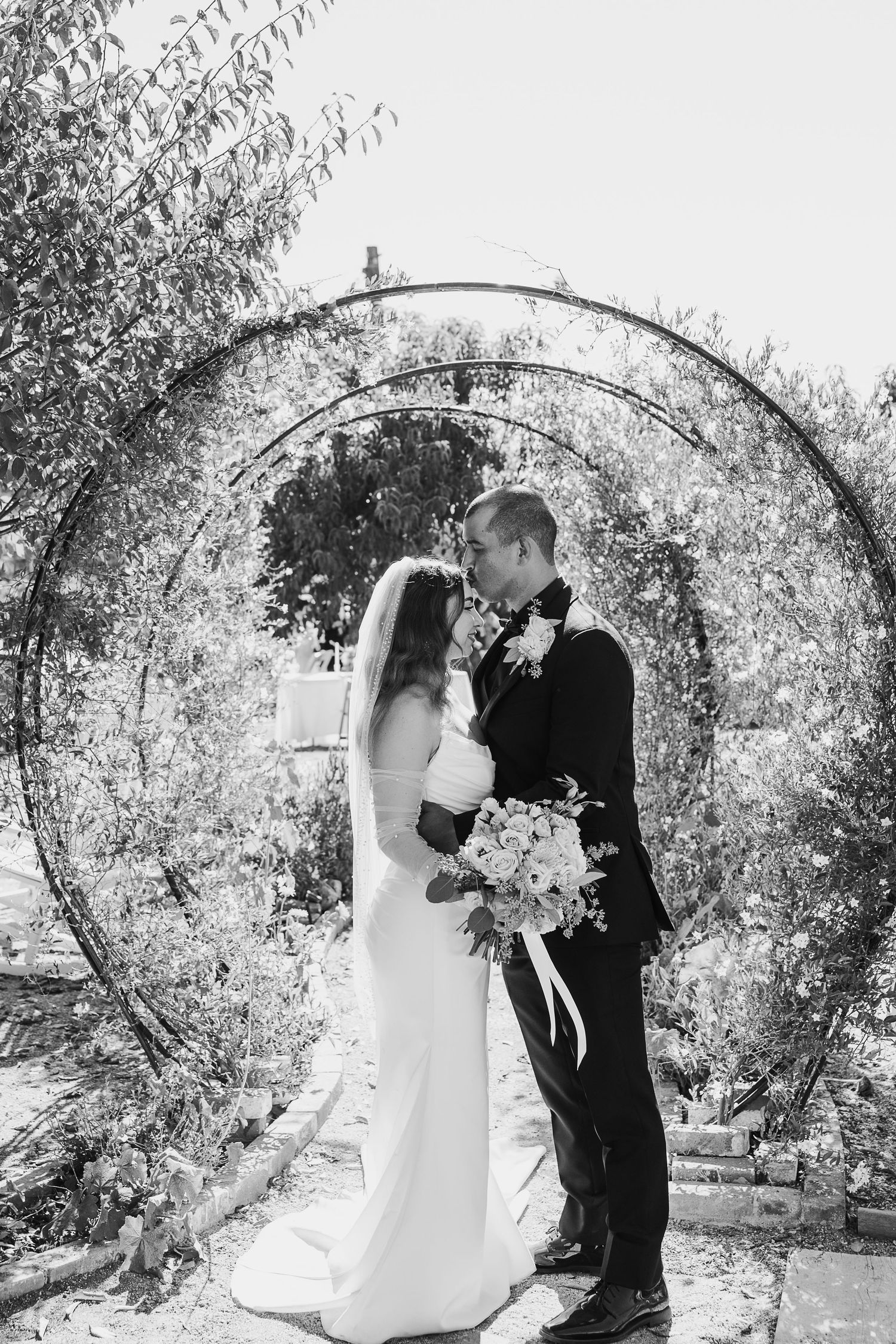 Black and white photo of wedding ceremony under floral arch in garden setting.