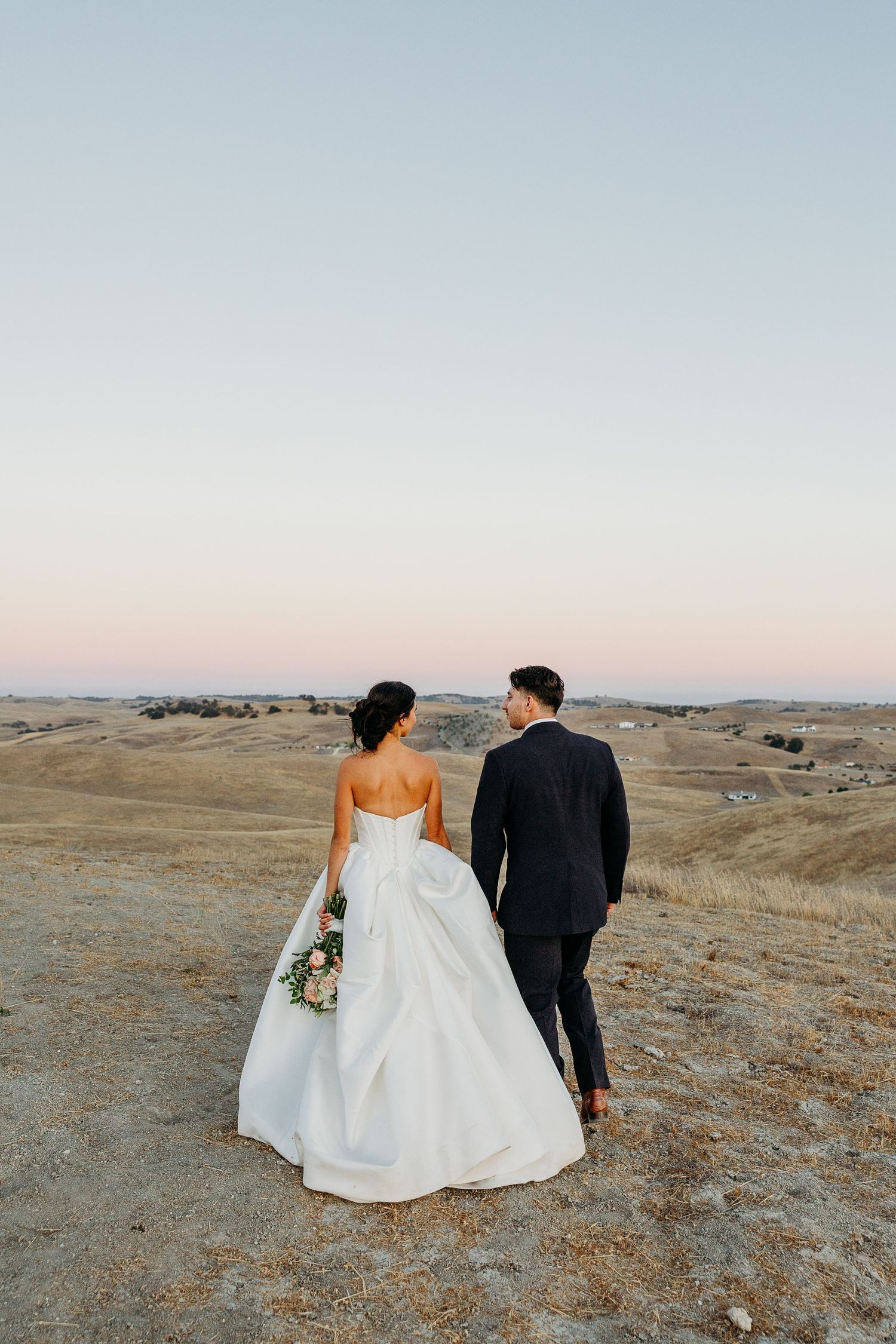 Newlyweds walking hand in hand across a scenic hilltop during golden hour.