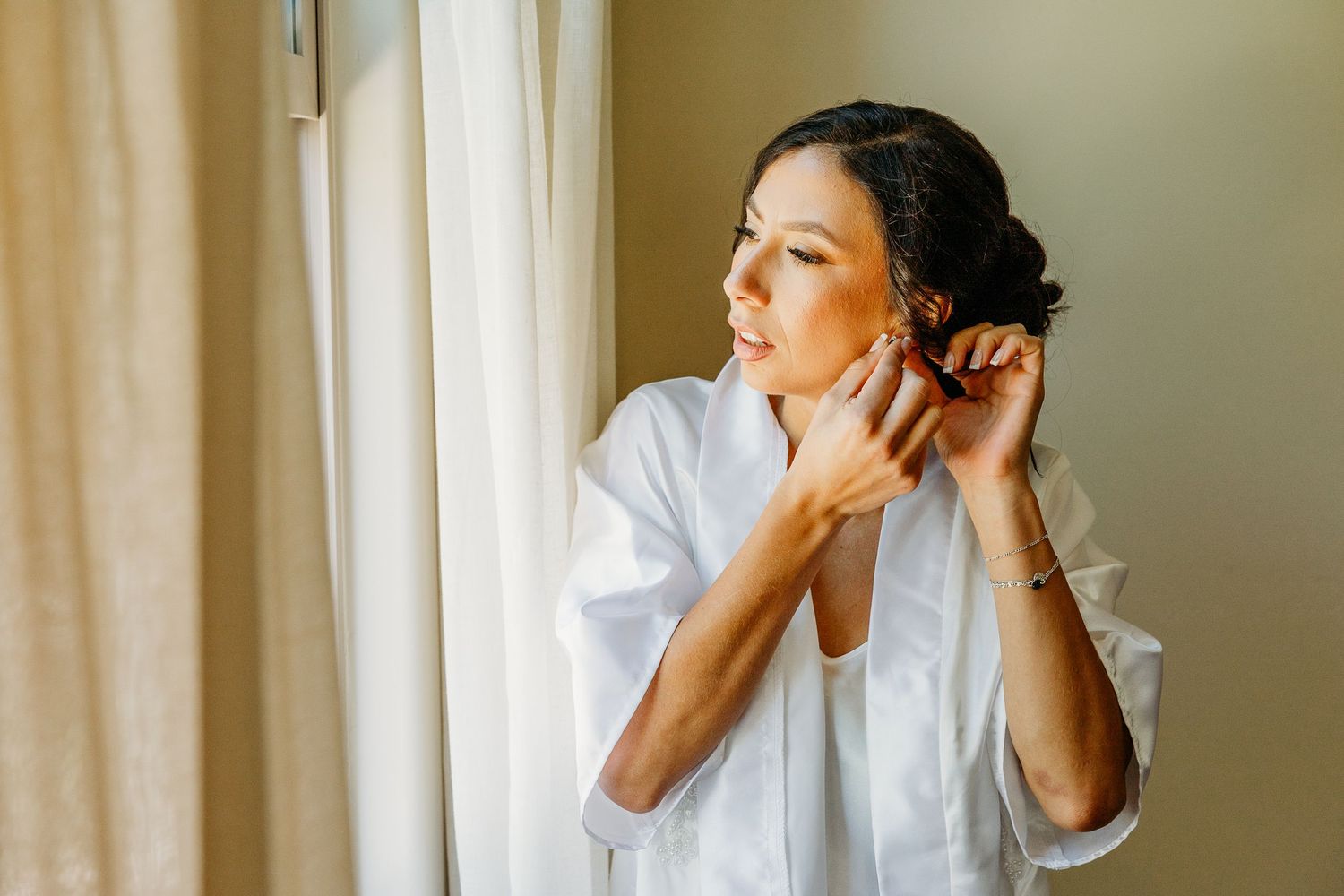 Person putting on earrings while standing near window in white robe during morning preparation.