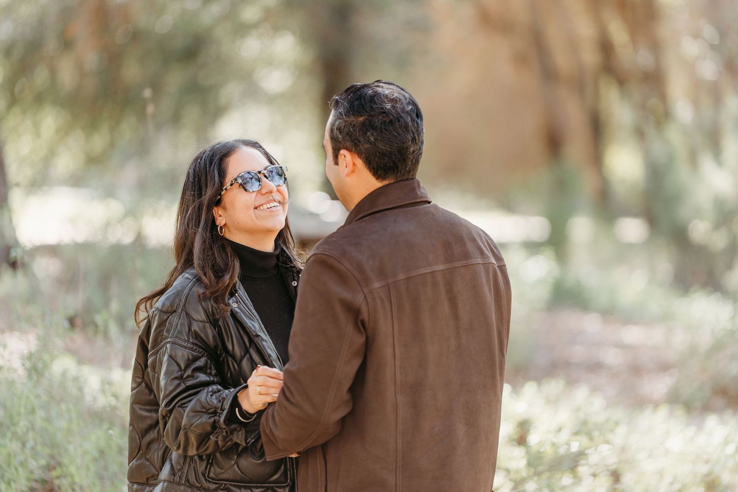 A couple shares a romantic moment outdoors in a natural setting with sunlit trees in the background.