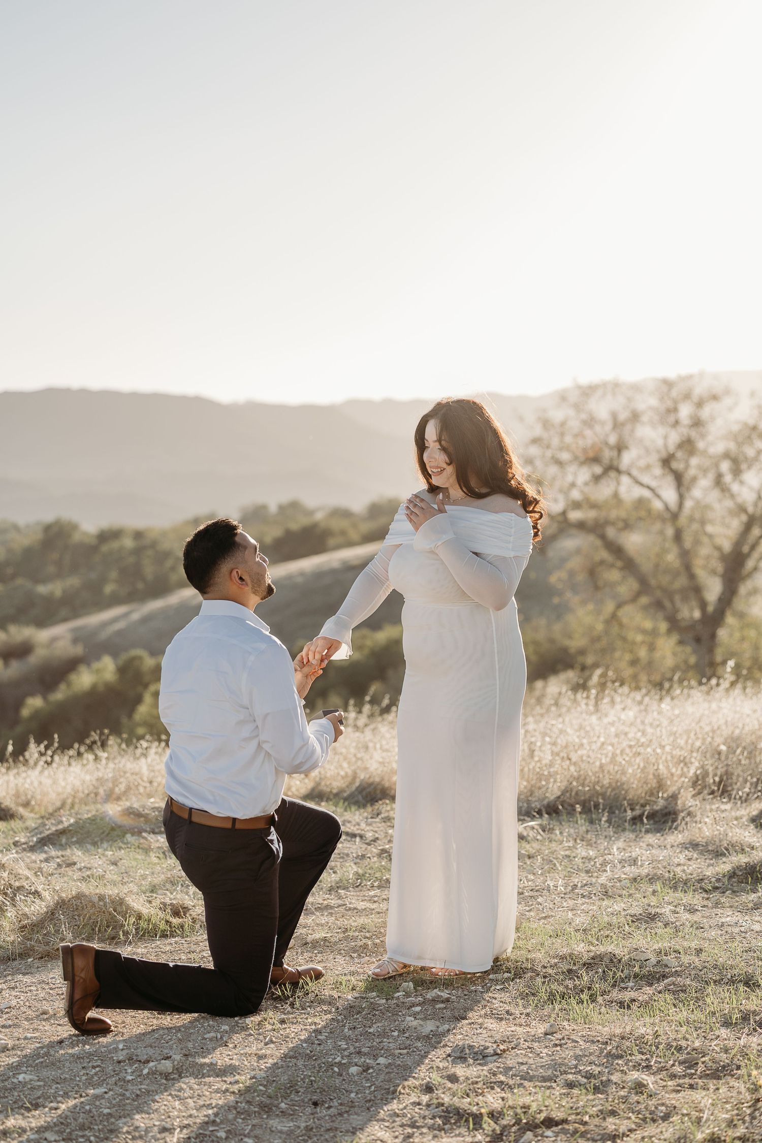 Surprise proposal on grassy hillside during golden hour sunset.