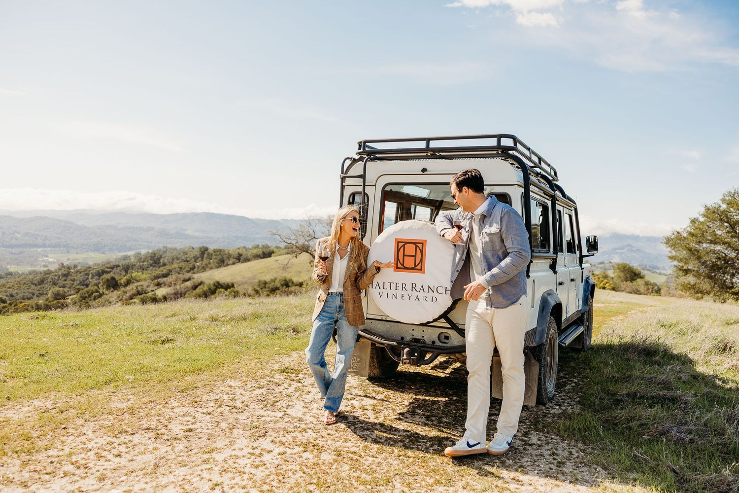Adventure couple stands beside vintage Land Rover overlooking mountain landscape.