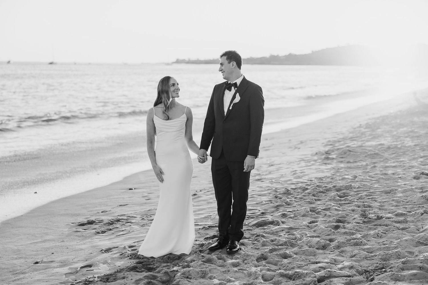 A couple walking hand in hand along a beach shoreline in formal wedding attire.