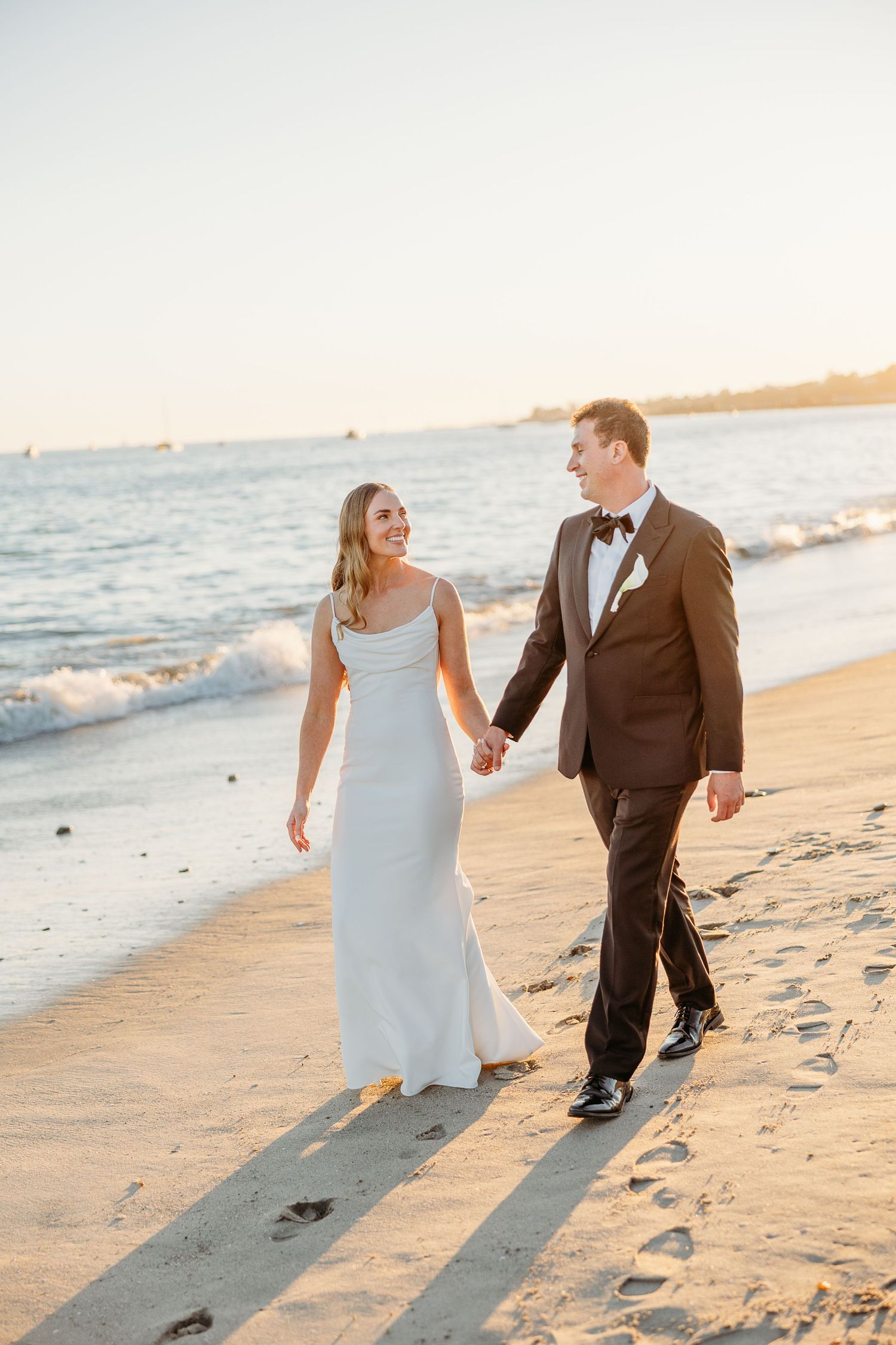 Newlyweds walk hand in hand along beach shoreline at sunset.