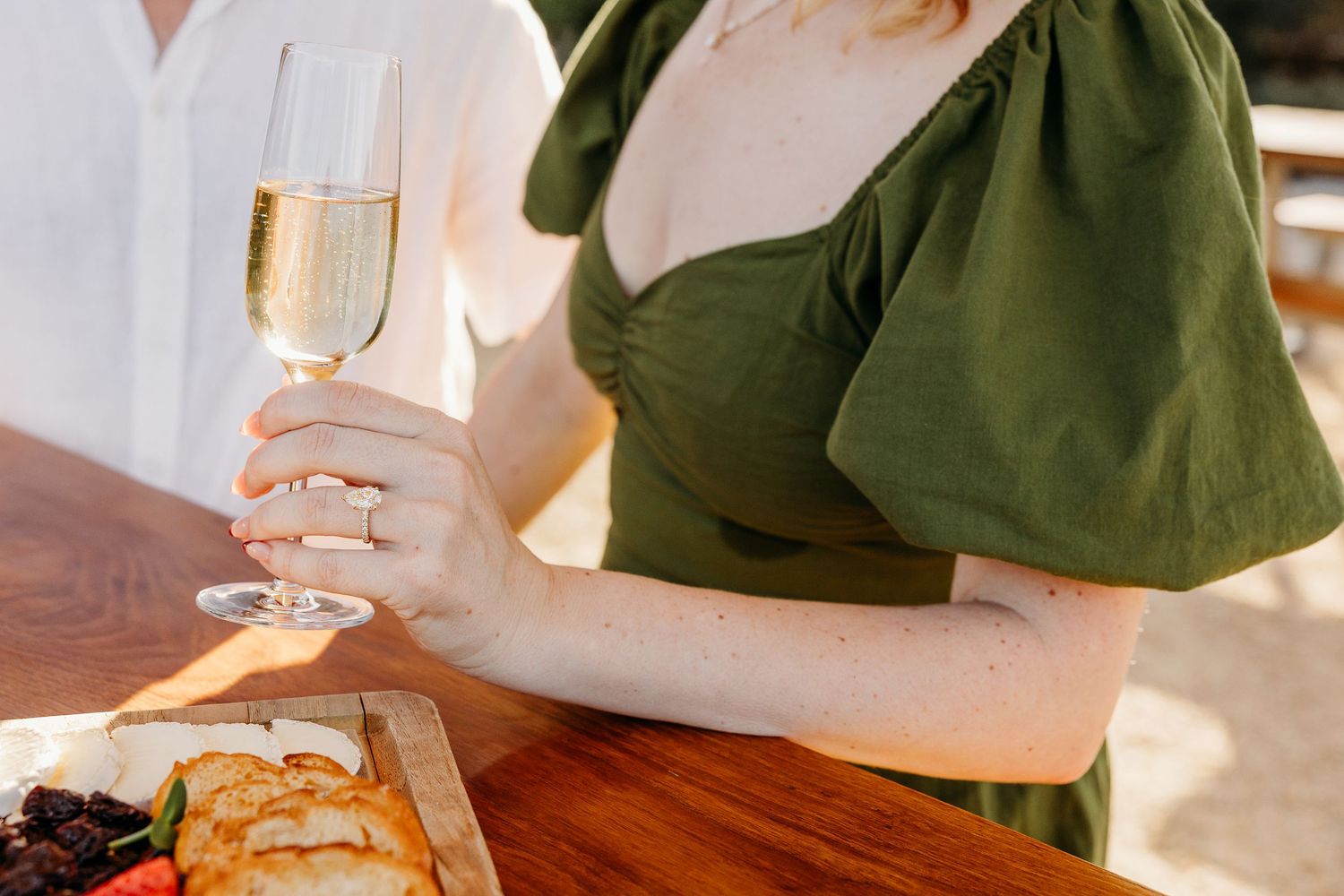 Person in a green dress holds a champagne glass at a wooden table with appetizers.