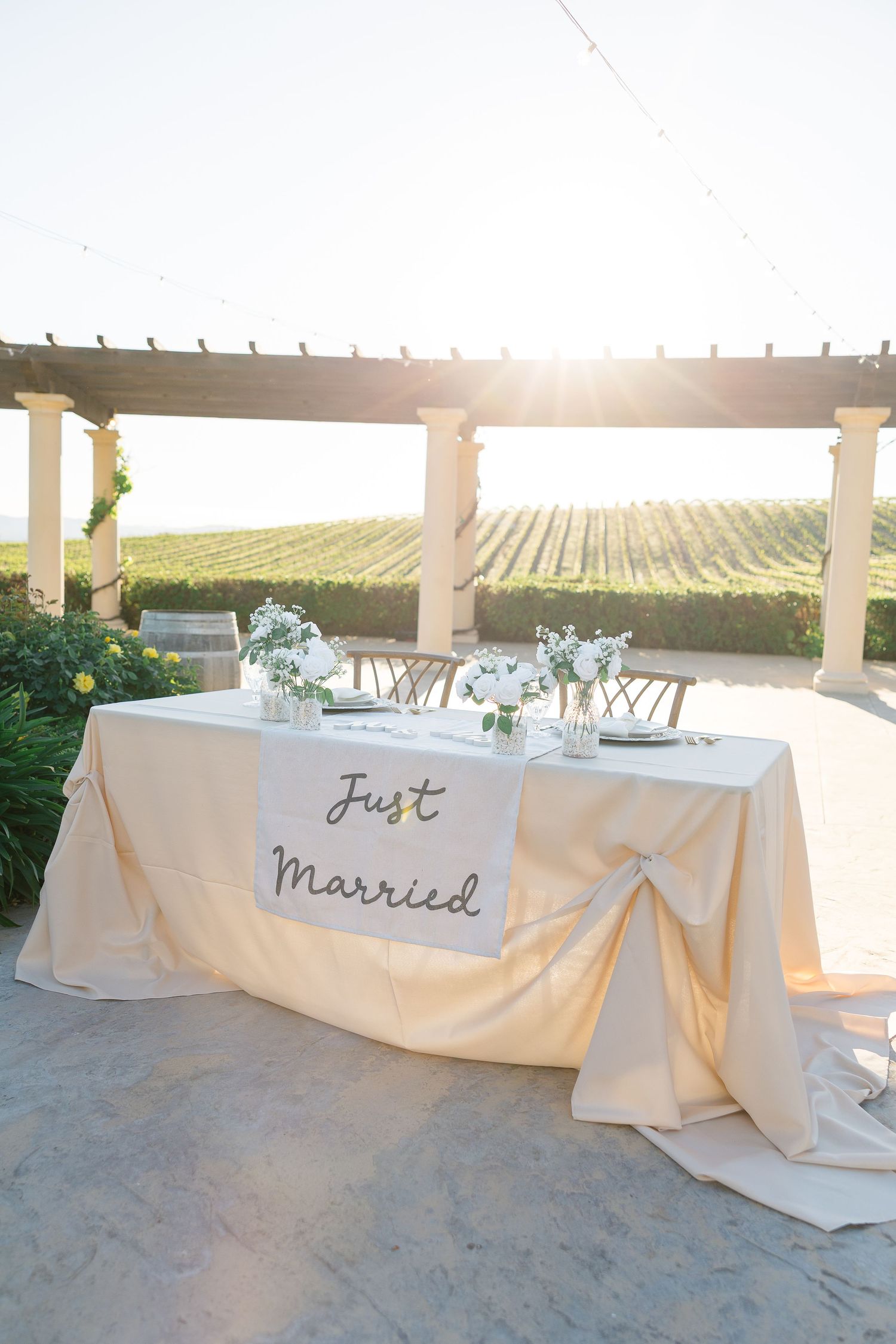 Just Married sweetheart table with white floral arrangements at sunset vineyard wedding reception.
