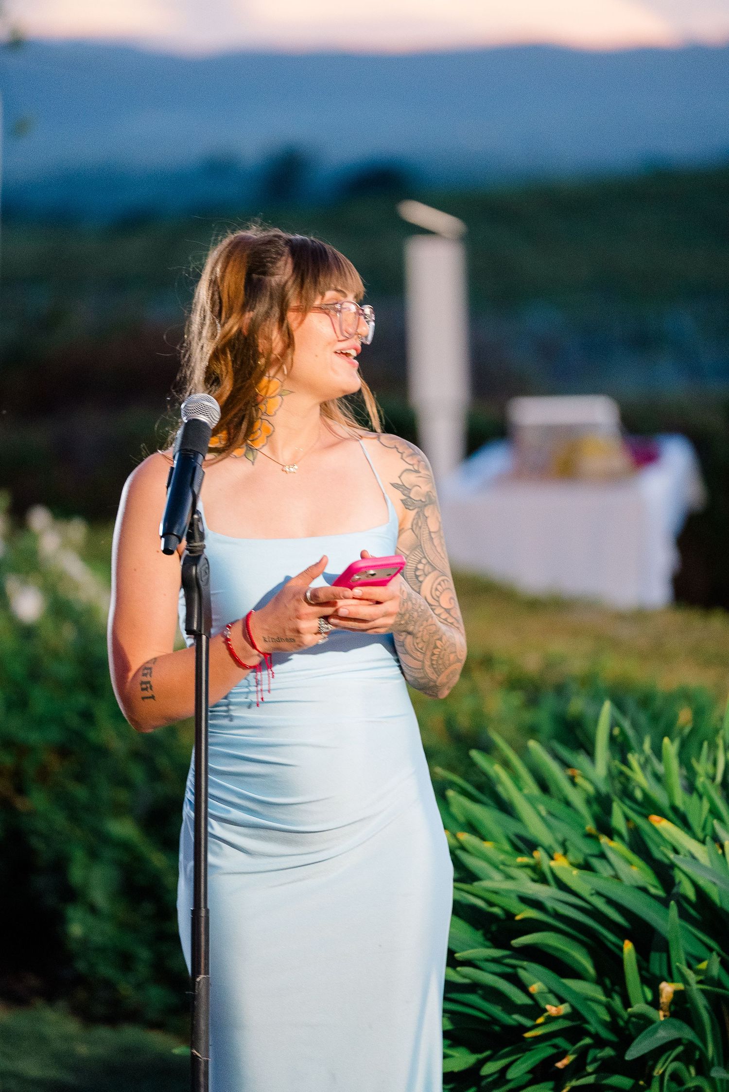 Tattooed woman in light blue gown gives speech at outdoor wedding reception at sunset.