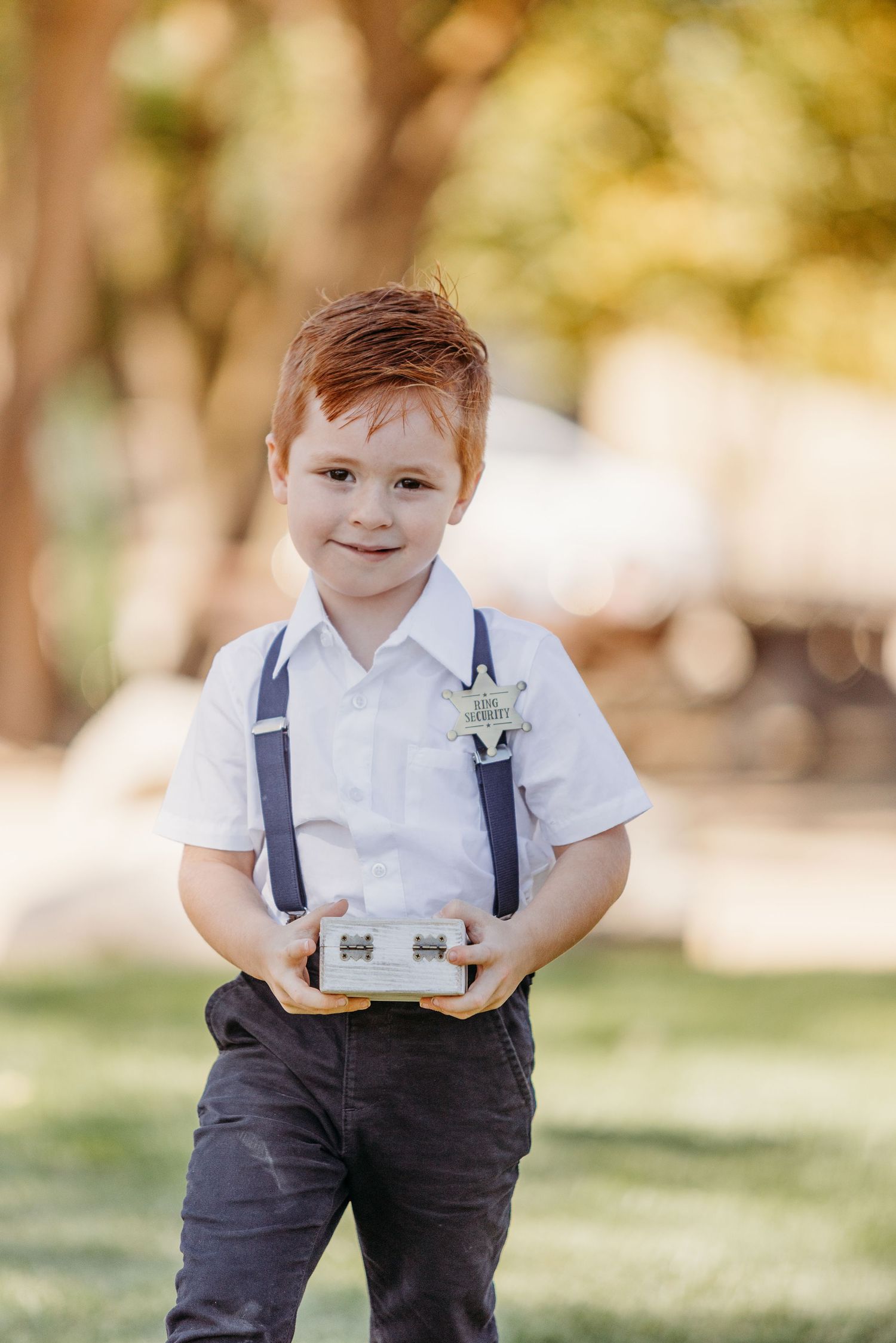 A small ring bearer in suspenders and dress pants stands outdoors against a blurred natural background.